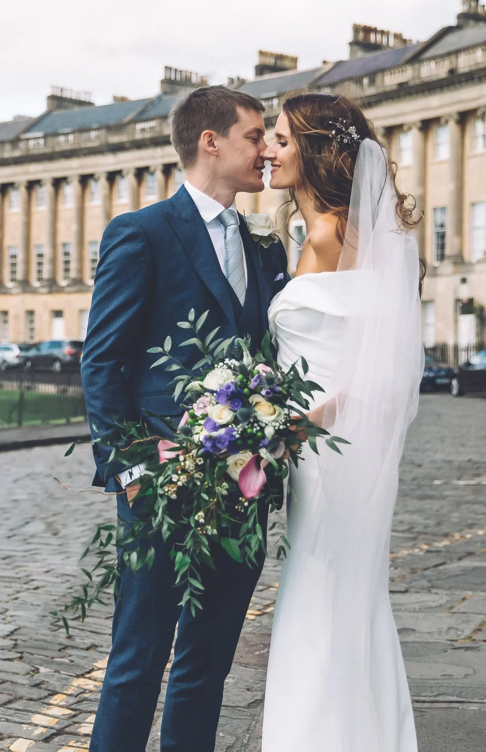 A wedding couple stands close together outdoors on a cobblestone street, with a historic building in the background. The groom wears a dark blue suit, white shirt, and light gray tie, holding a bouquet of flowers. The bride wears a white wedding gown