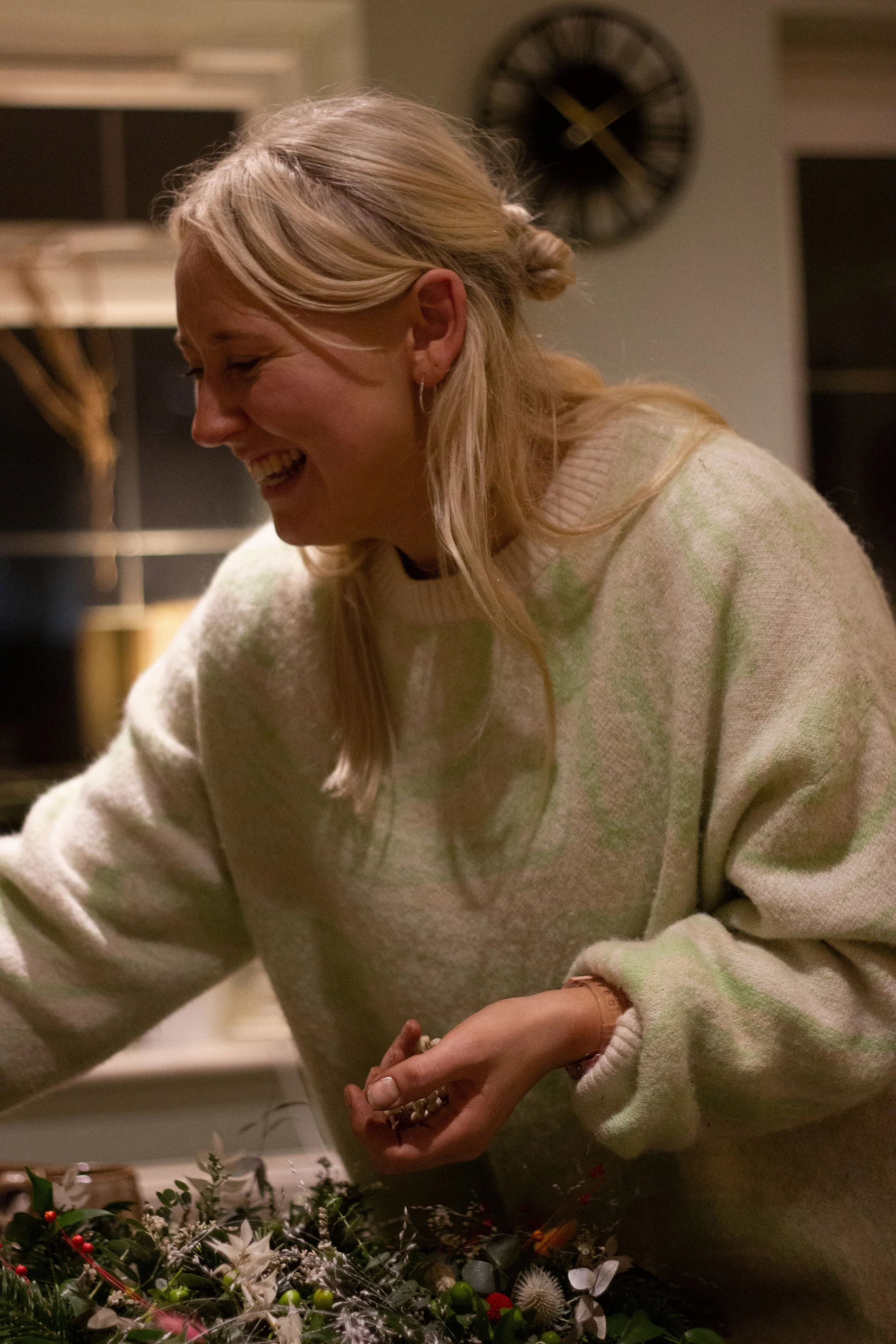 A woman with blonde hair in a loose braid, smiling and leaning over a table with Christmas decorations, wearing a cream-colored sweater.