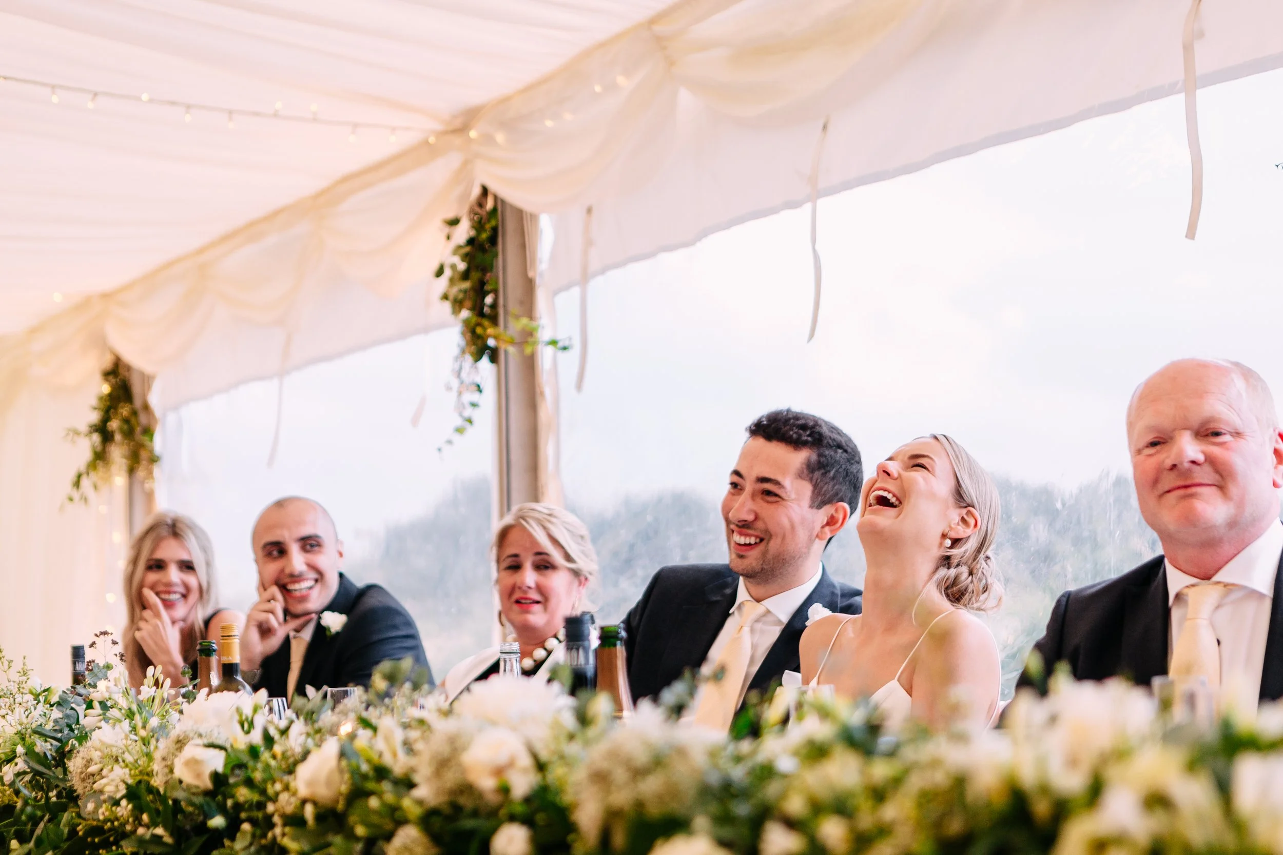 People at a wedding reception seated at a table with floral arrangements, all smiling and laughing inside a decorated tent.