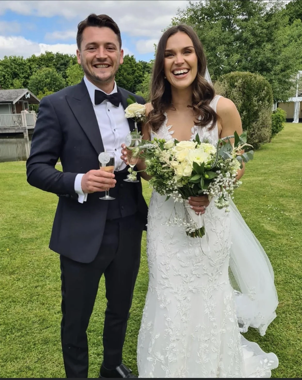 A happy bride and groom standing outdoors on a grassy area, smiling at the camera, holding glasses of champagne. The groom is in a black tuxedo with a bow tie, and the bride is wearing a white wedding gown with floral lace details and holding a bouqu