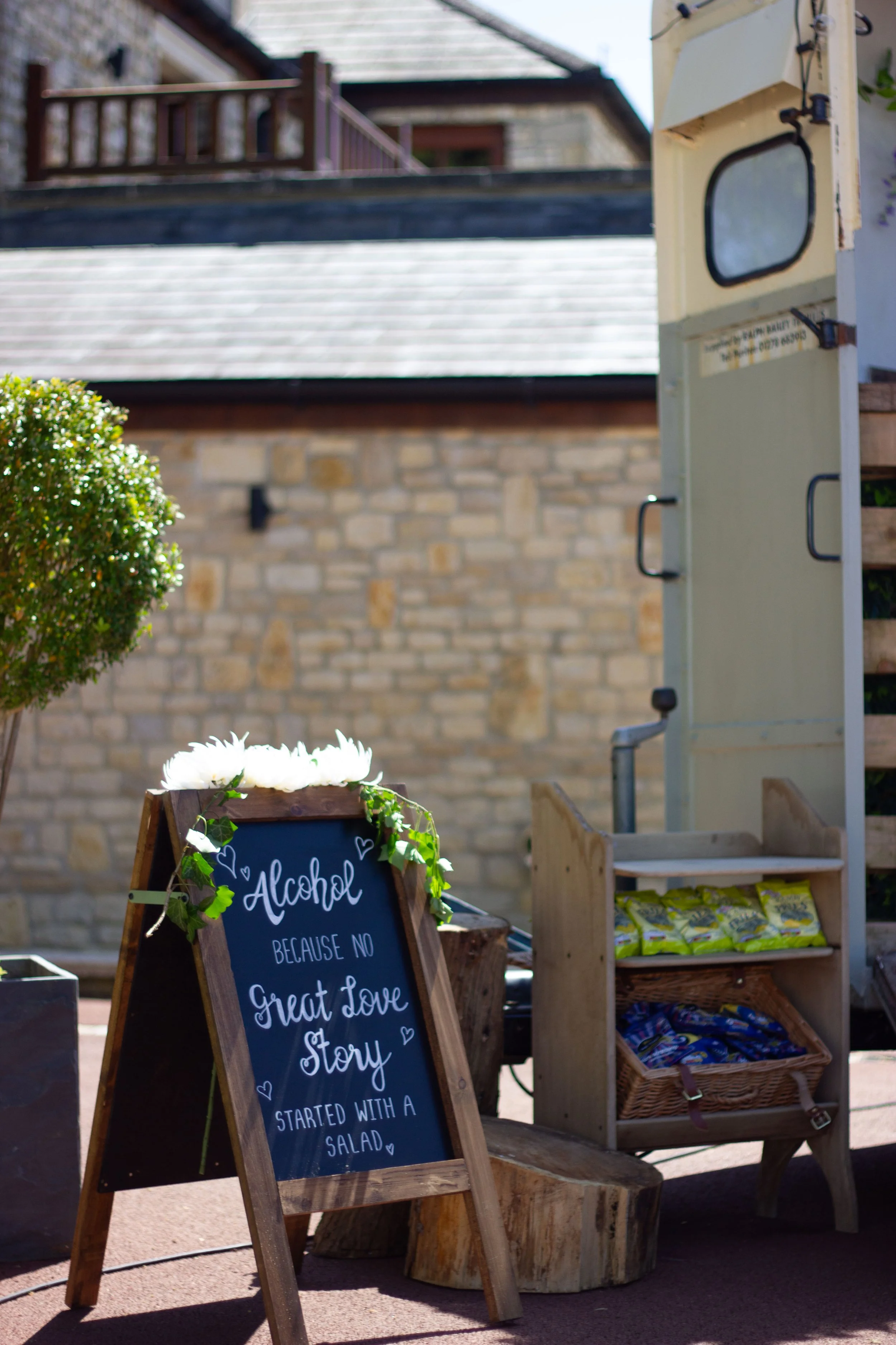 Outdoor scene with a chalkboard sign that reads, 'Alcohol because no great love story started with a salad,' surrounded by decorative greenery, a tree, and a vintage trailer, with a brick building in the background.