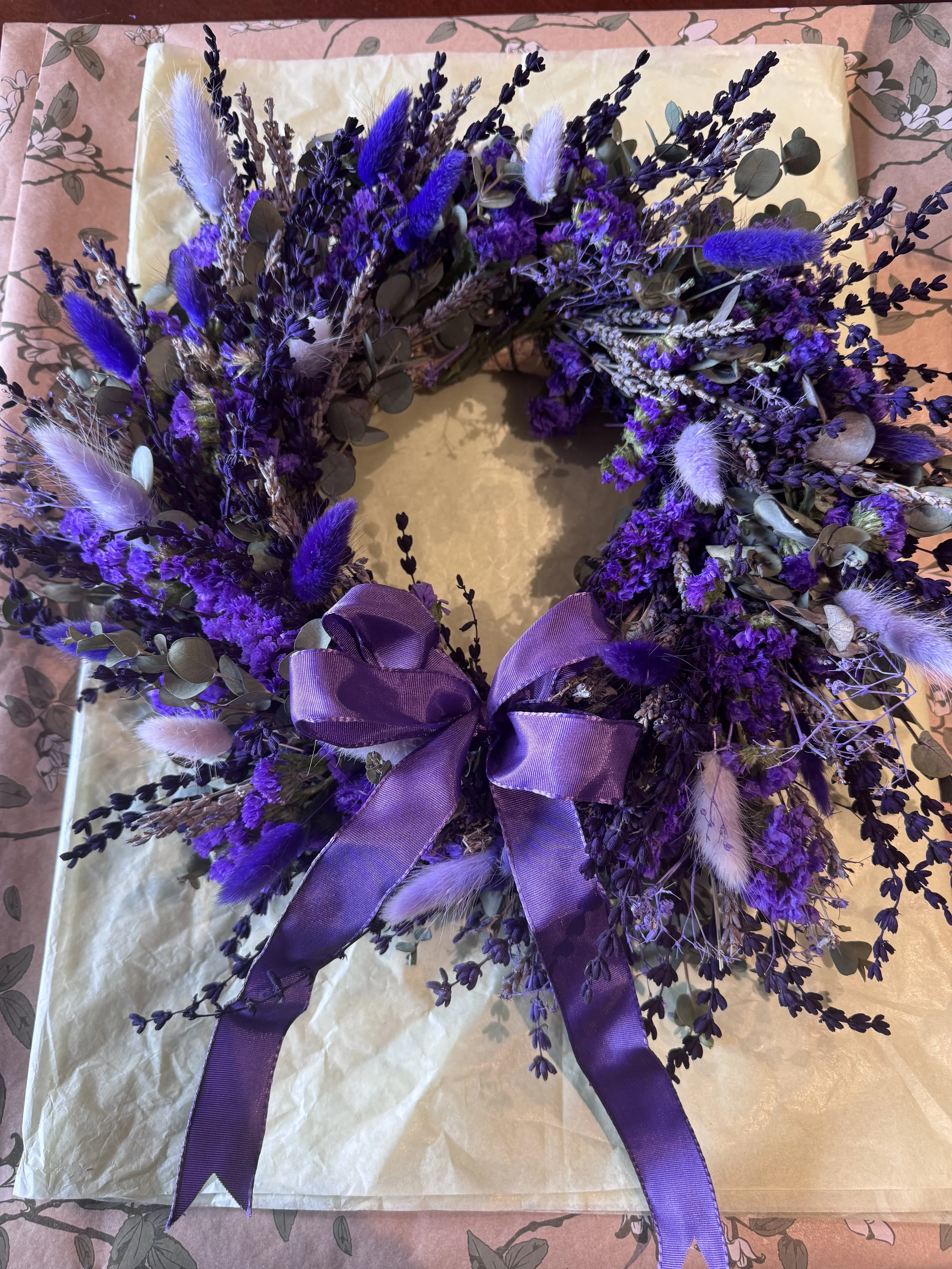 A purple floral wreath with a purple ribbon bow, placed on wrapping paper with a floral pattern.