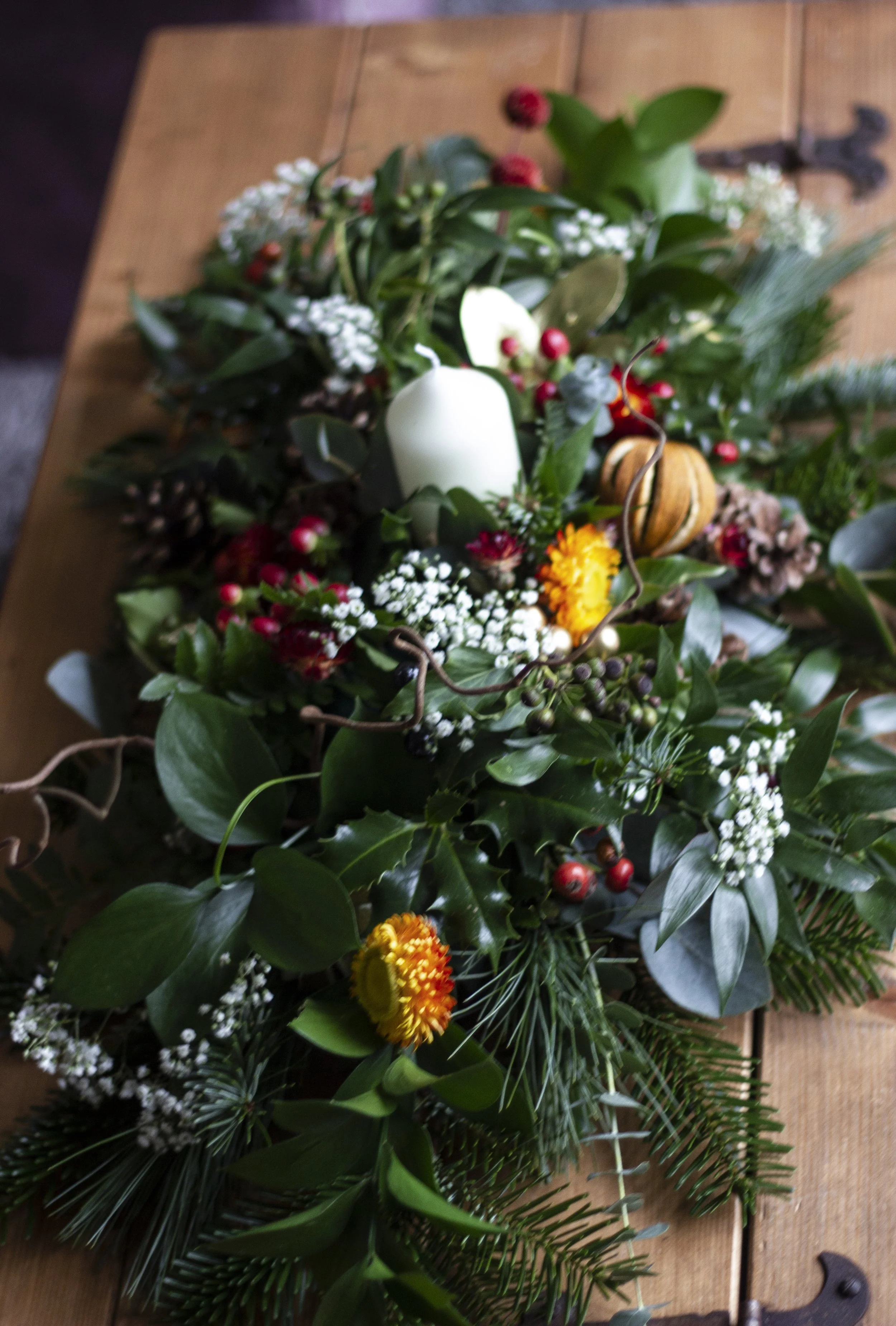 A festive Christmas arrangement on a wooden table, featuring green foliage, white candles, red berries, pinecones, yellow and orange flowers, and a small pumpkin.