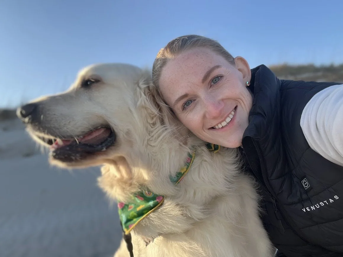 A smiling woman taking a selfie with her golden retriever wearing a colorful bandana outside on a clear day.
