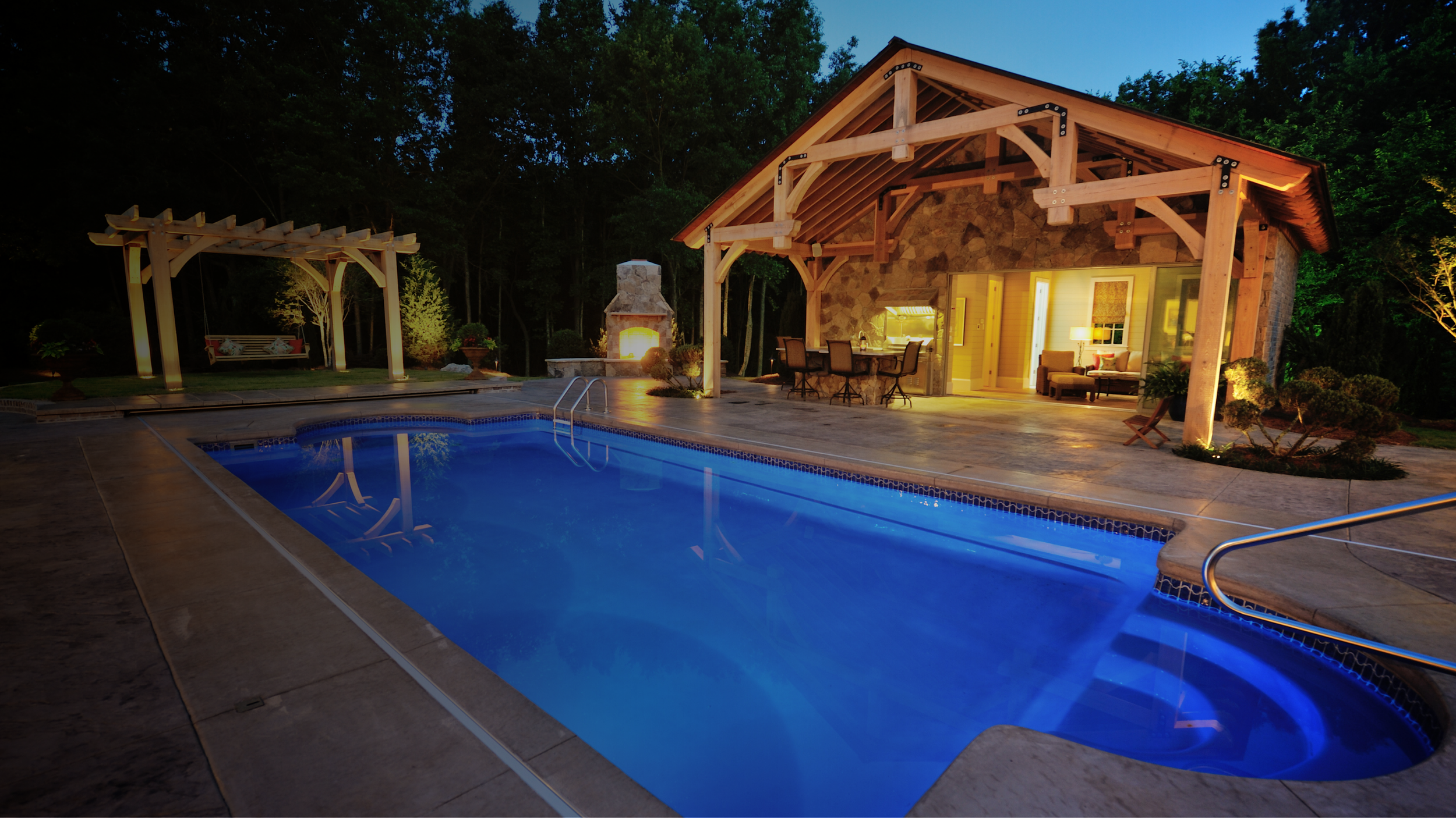 Nighttime view of a backyard with a lit swimming pool, a stone house with a covered patio, and a firepit area with a swing and outdoor seating.