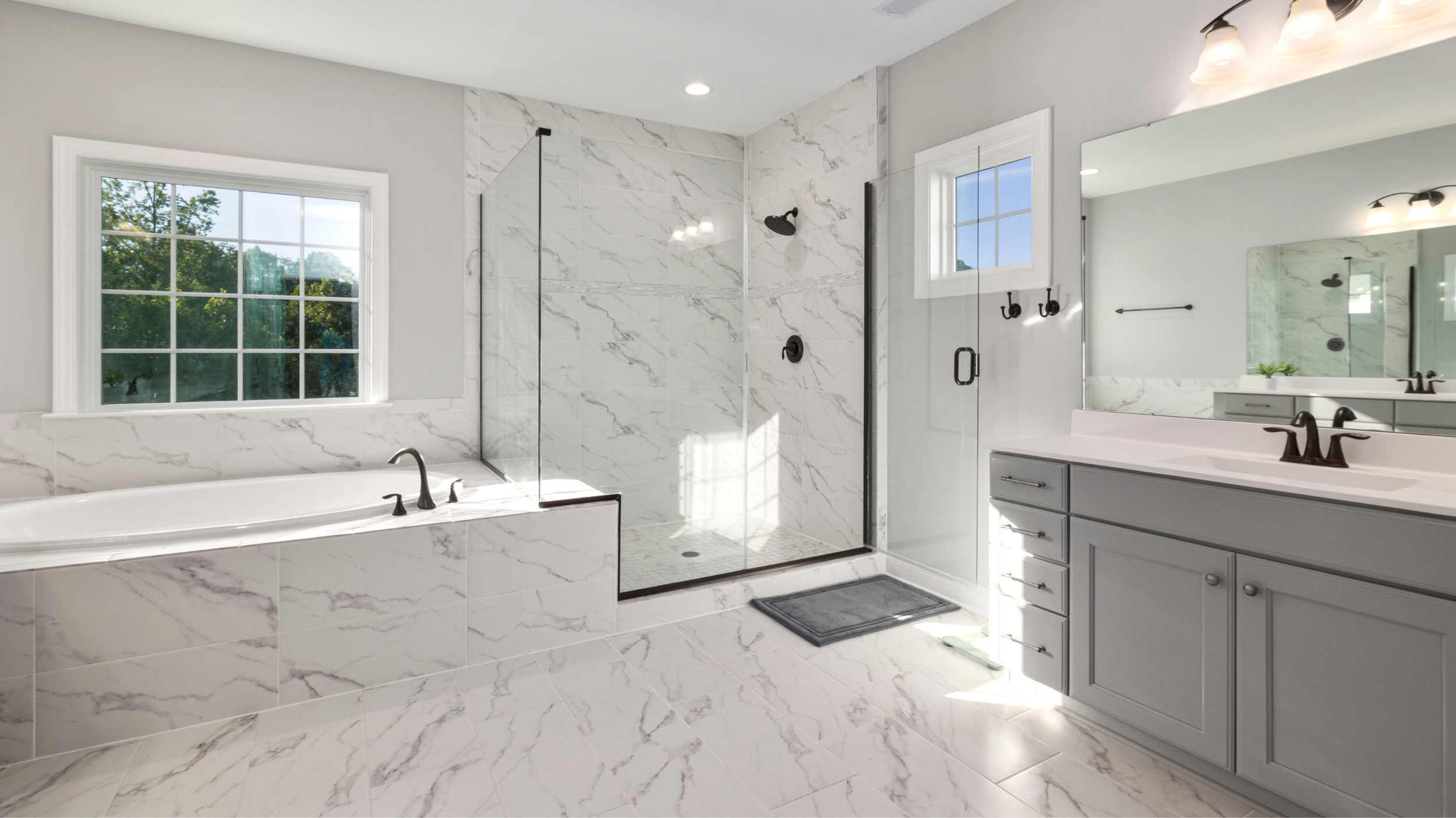 Modern bathroom with marble tiles, a bathtub, a glass-enclosed shower, a window, and a light gray vanity with a large mirror.
