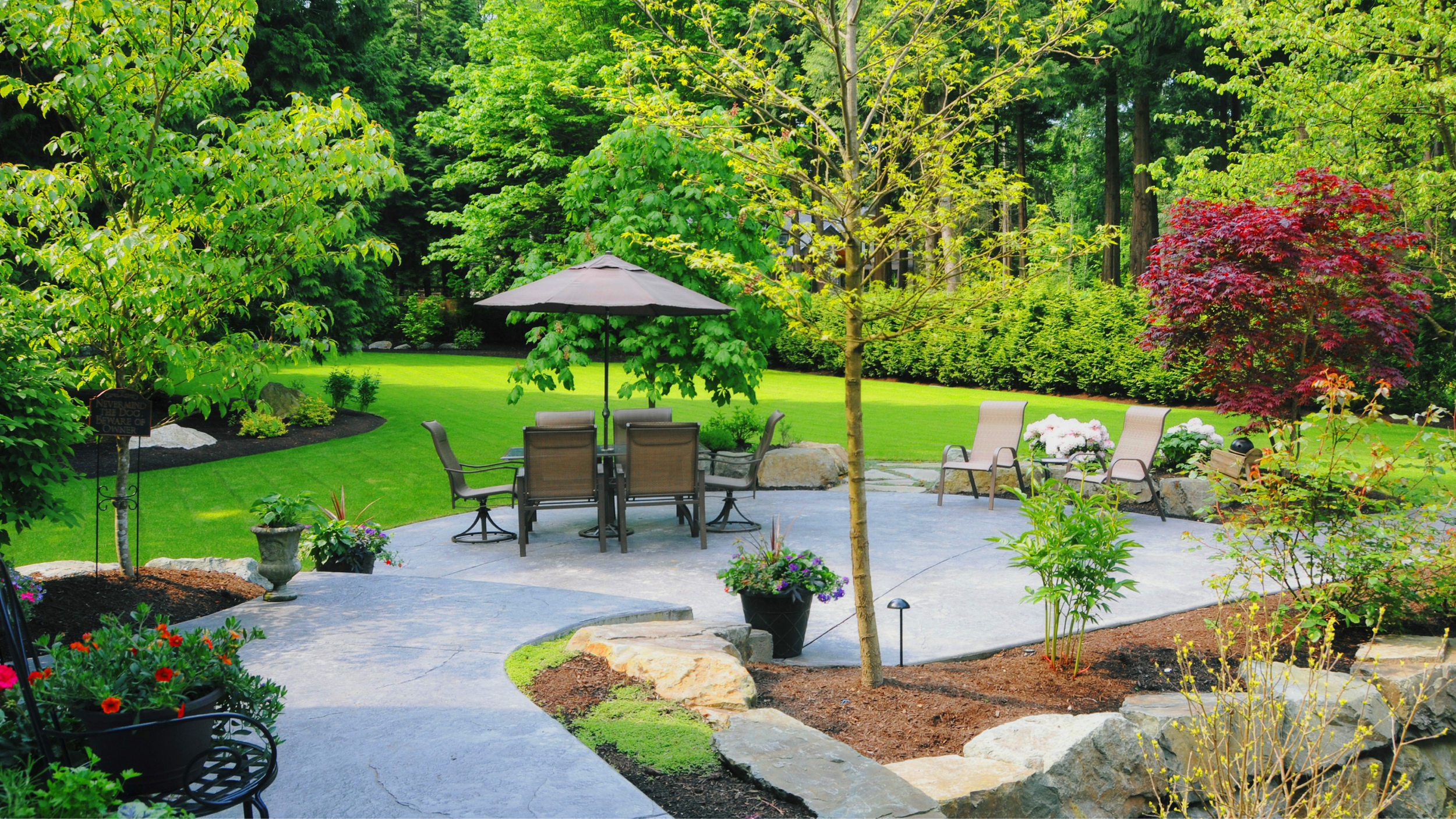 A landscaped backyard patio with outdoor furniture, including a table with an umbrella, surrounded by green trees, plants, and lawn, with flower pots and a red-leafed tree in the background.