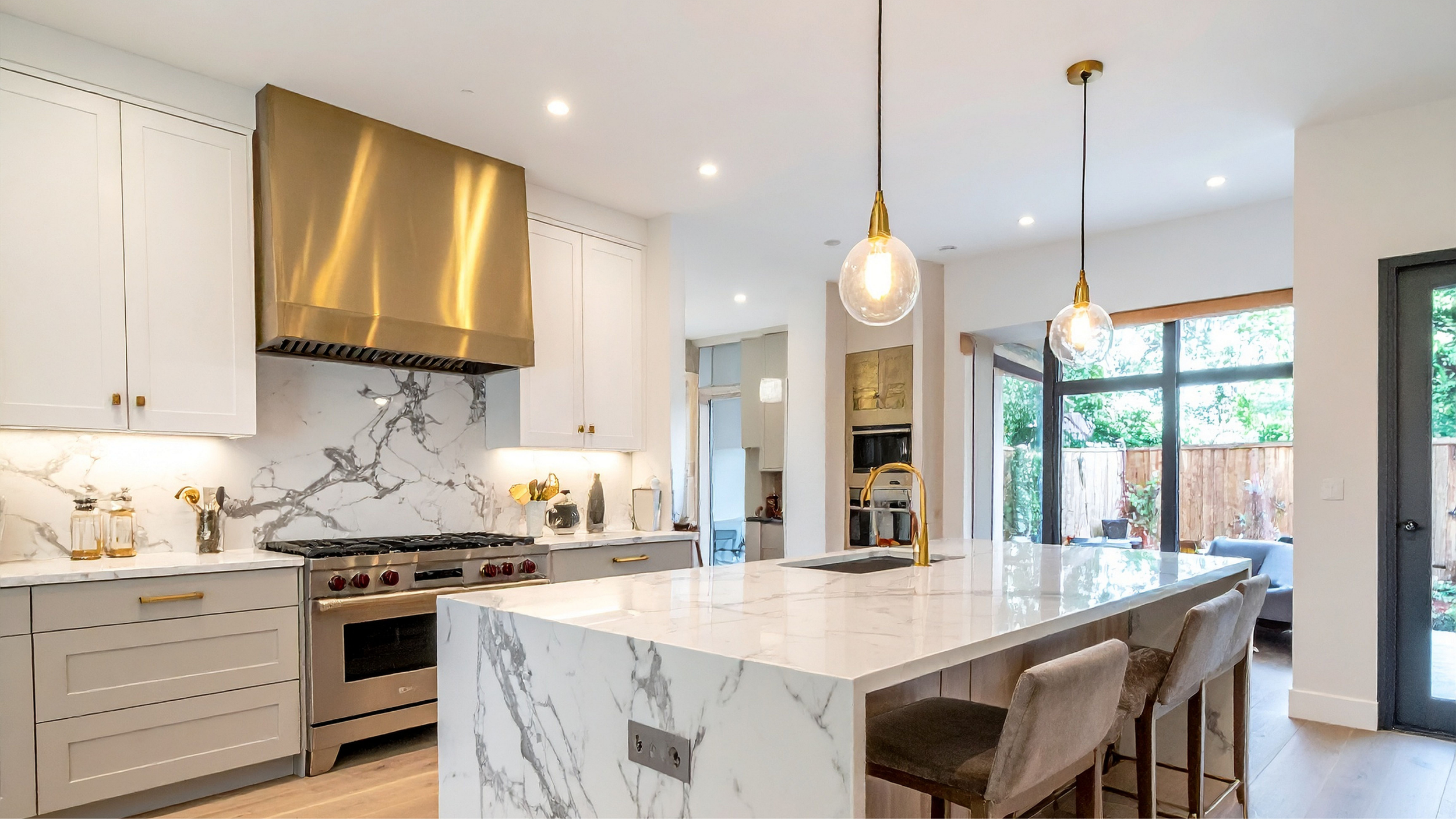 Modern kitchen with white cabinetry, marble backsplash and countertops, a stainless steel range with a large bronze range hood, pendant lights, and a large marble island with seating. Large windows and a door lead to an outdoor patio.