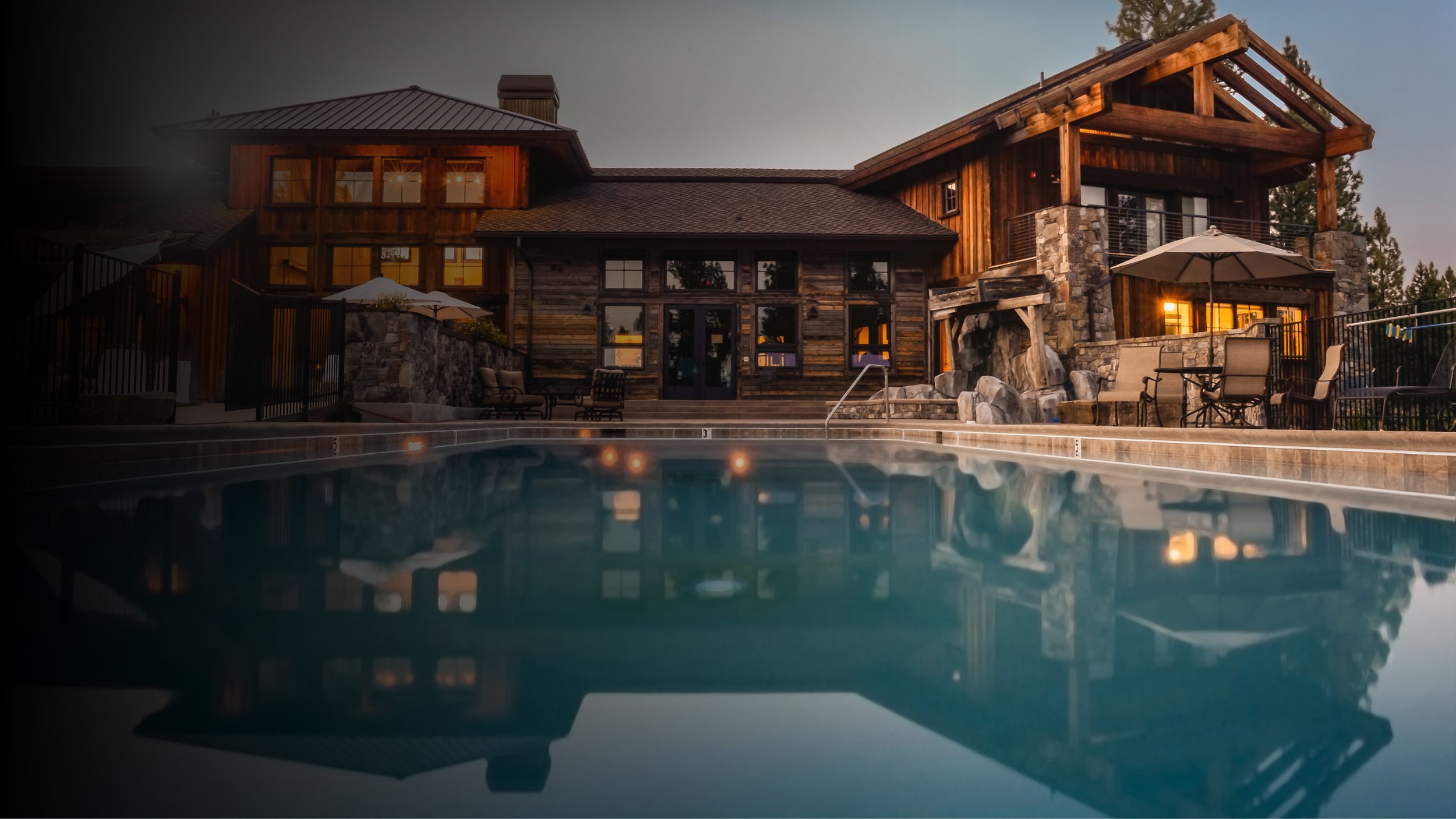 A large house with wooden and stone exterior, lit warmly from inside, seen from the edge of a swimming pool at dusk. Outdoor furniture and umbrellas are visible on the patio.