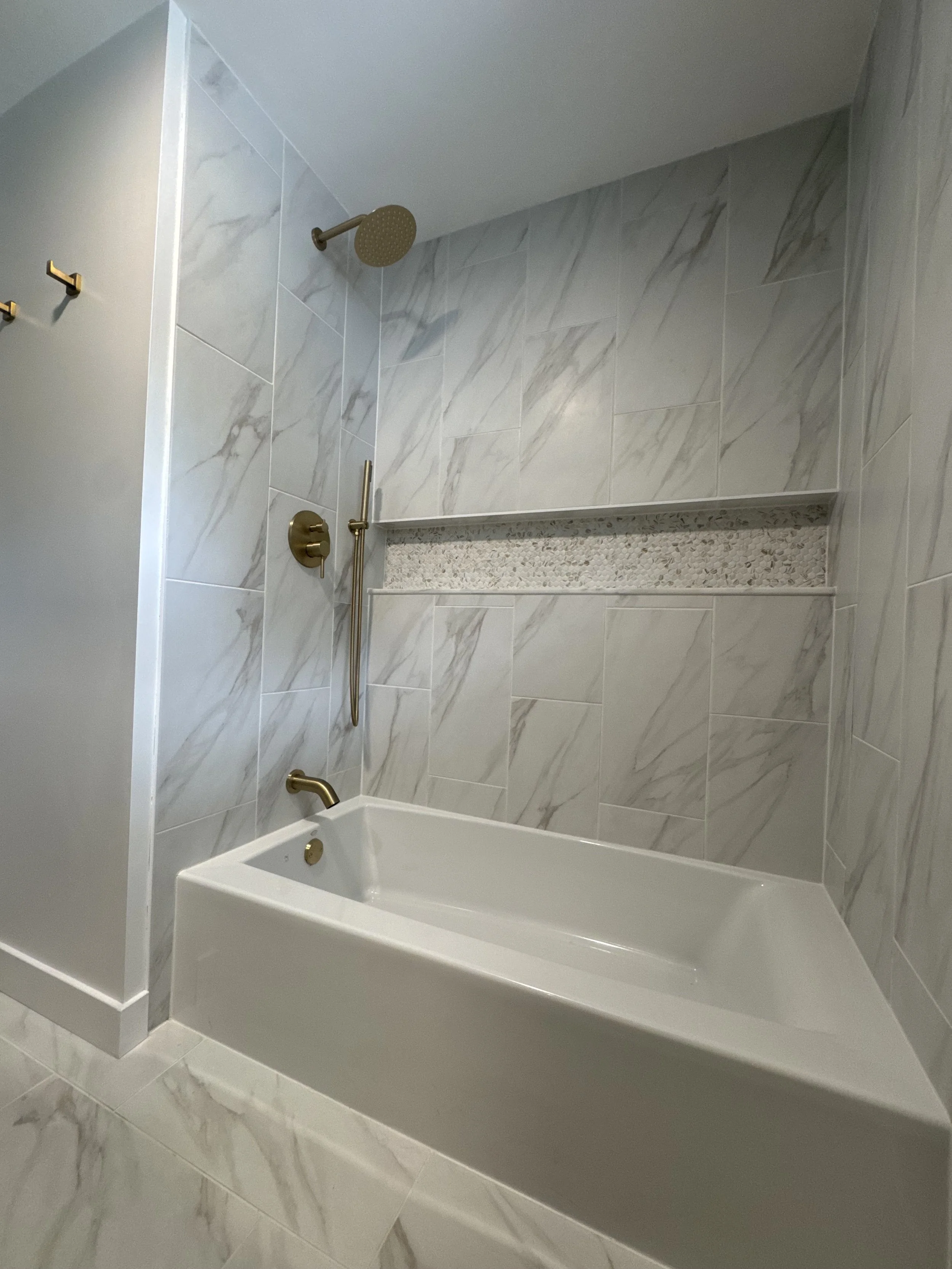 Bathroom with a white bathtub, marble tiled wall, and gold fixtures including a showerhead, faucet, and a handheld shower wand.