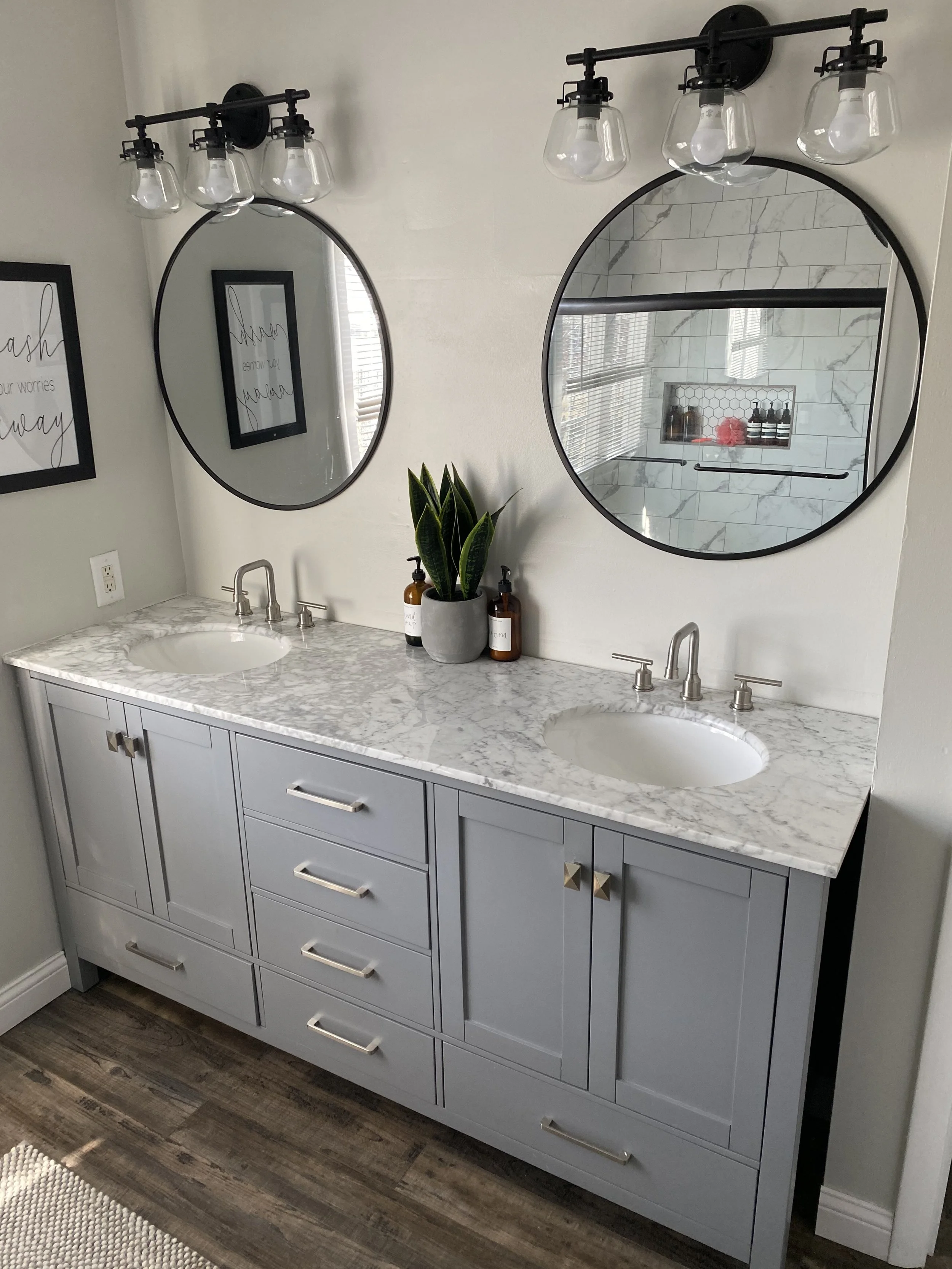 Gray double vanity with marble countertop, two oval mirrors, black light fixtures with exposed bulbs, a potted plant, and brown bottles, in a modern bathroom.