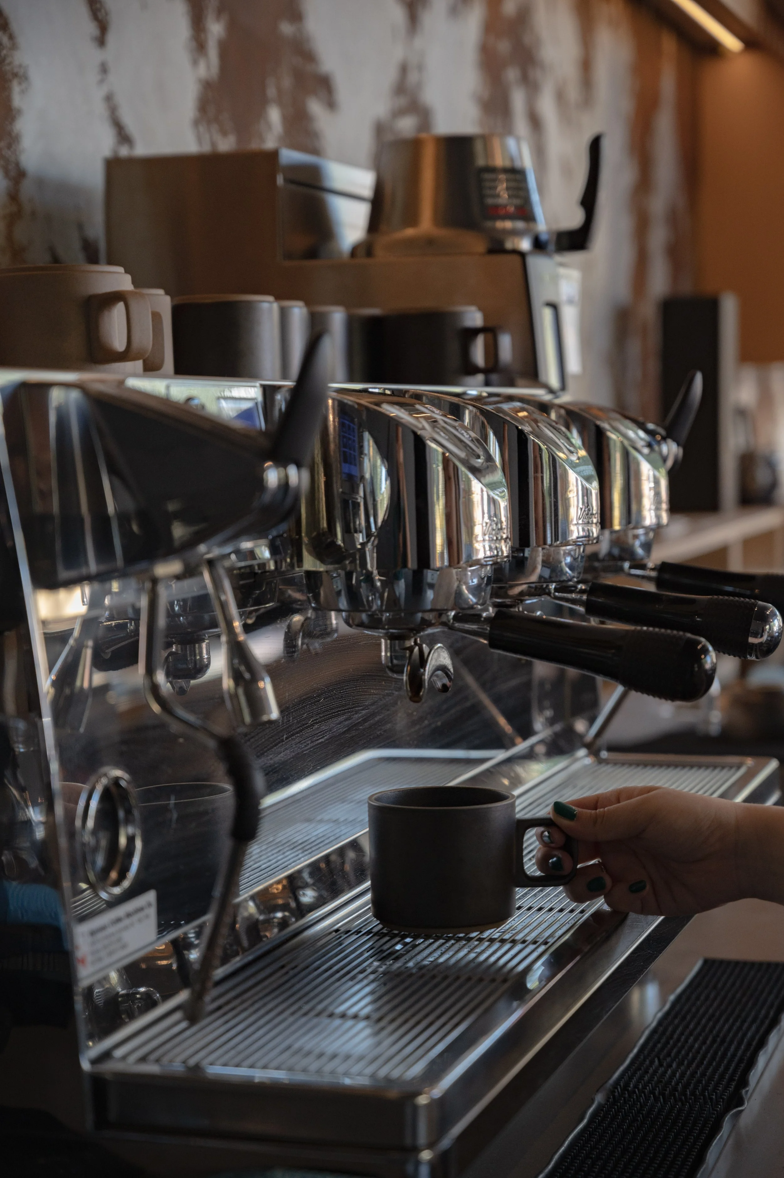 A person holding a black coffee mug under an espresso machine in a coffee shop.