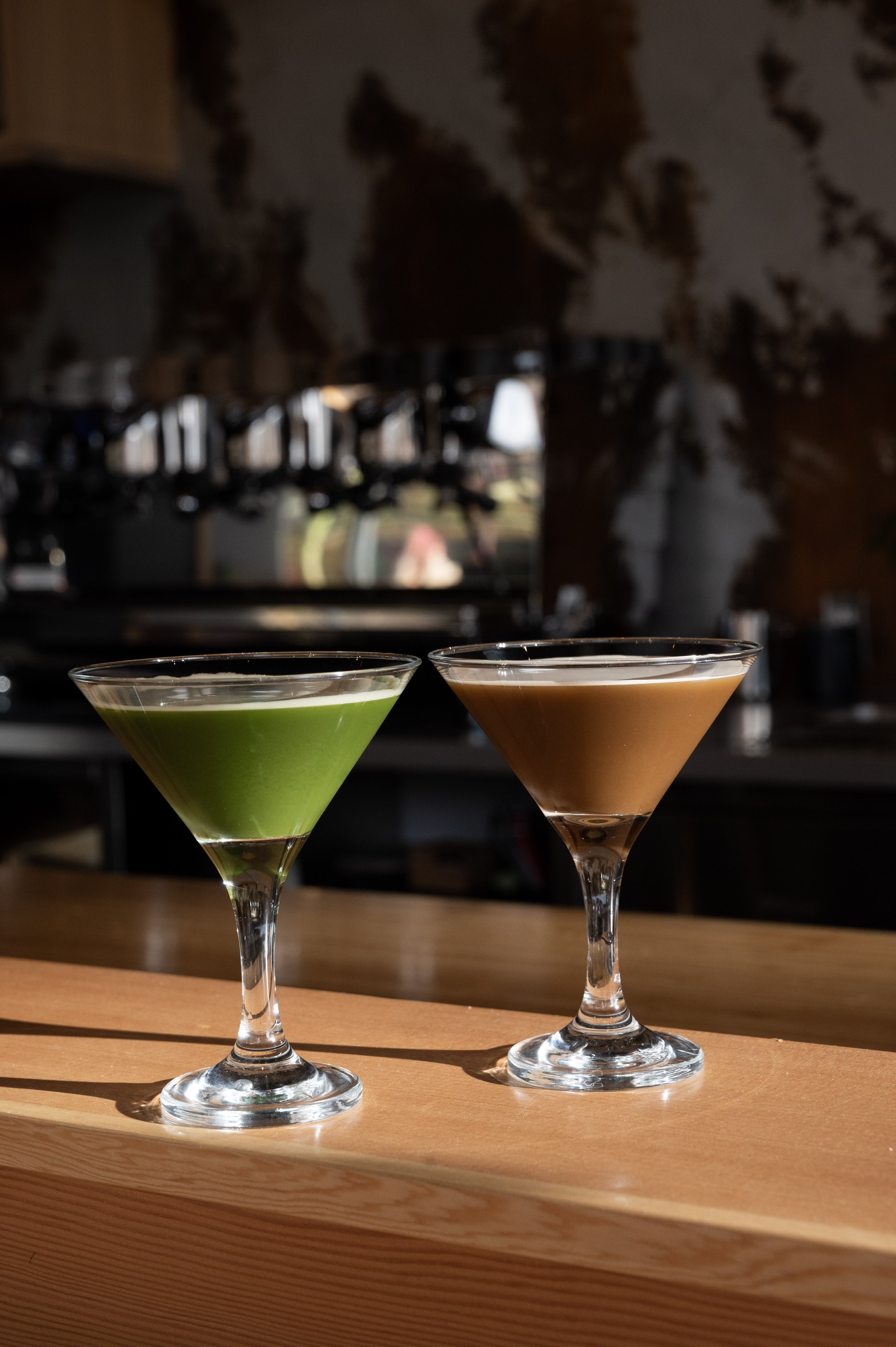 Two martini glasses, one with a green cocktail and one with a brown cocktail, placed on a wooden surface in a bar setting.