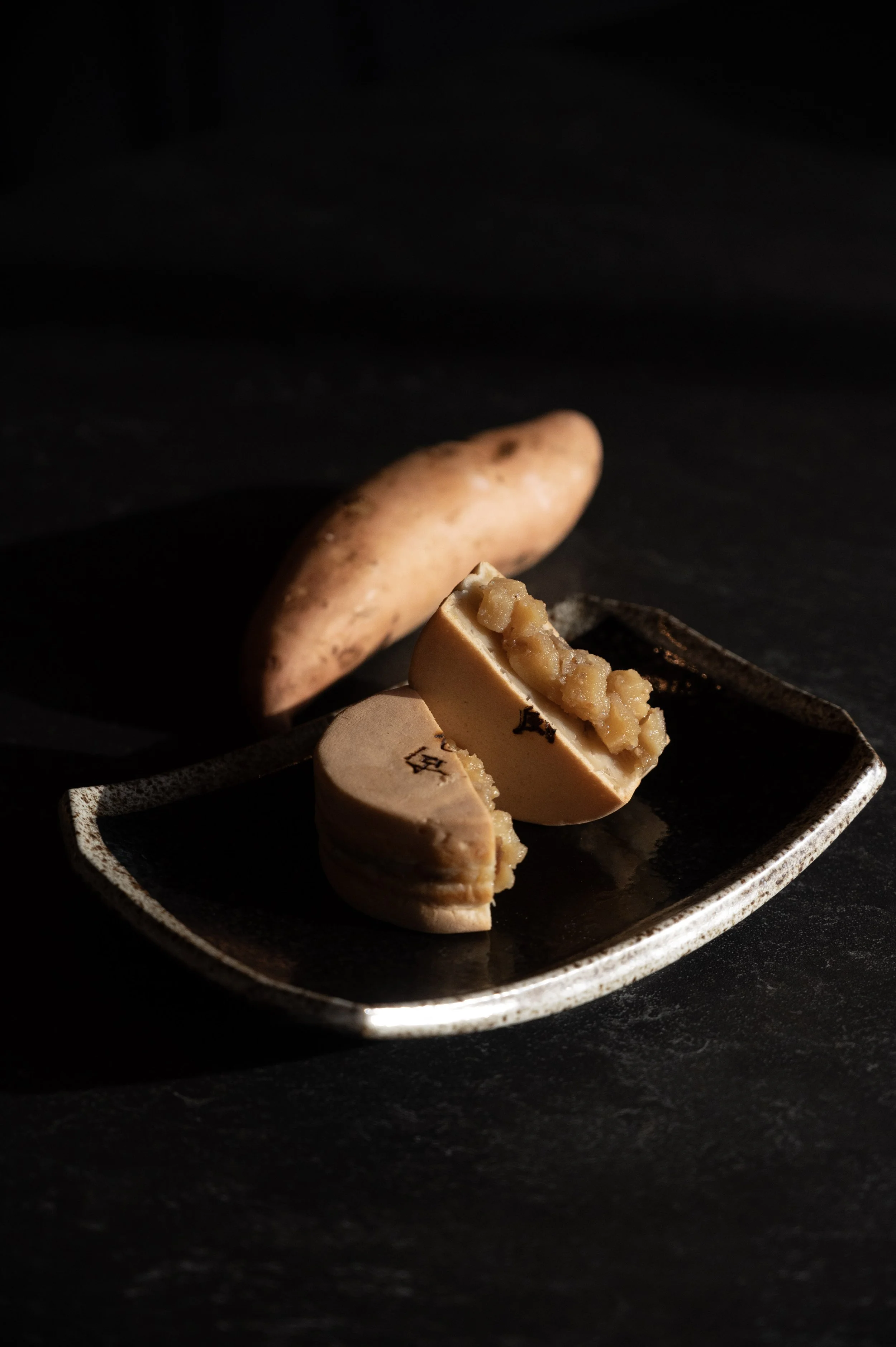 A plate with two pieces of steamed bun filled with mashed sweet potato, with a whole sweet potato in the background on a dark surface.