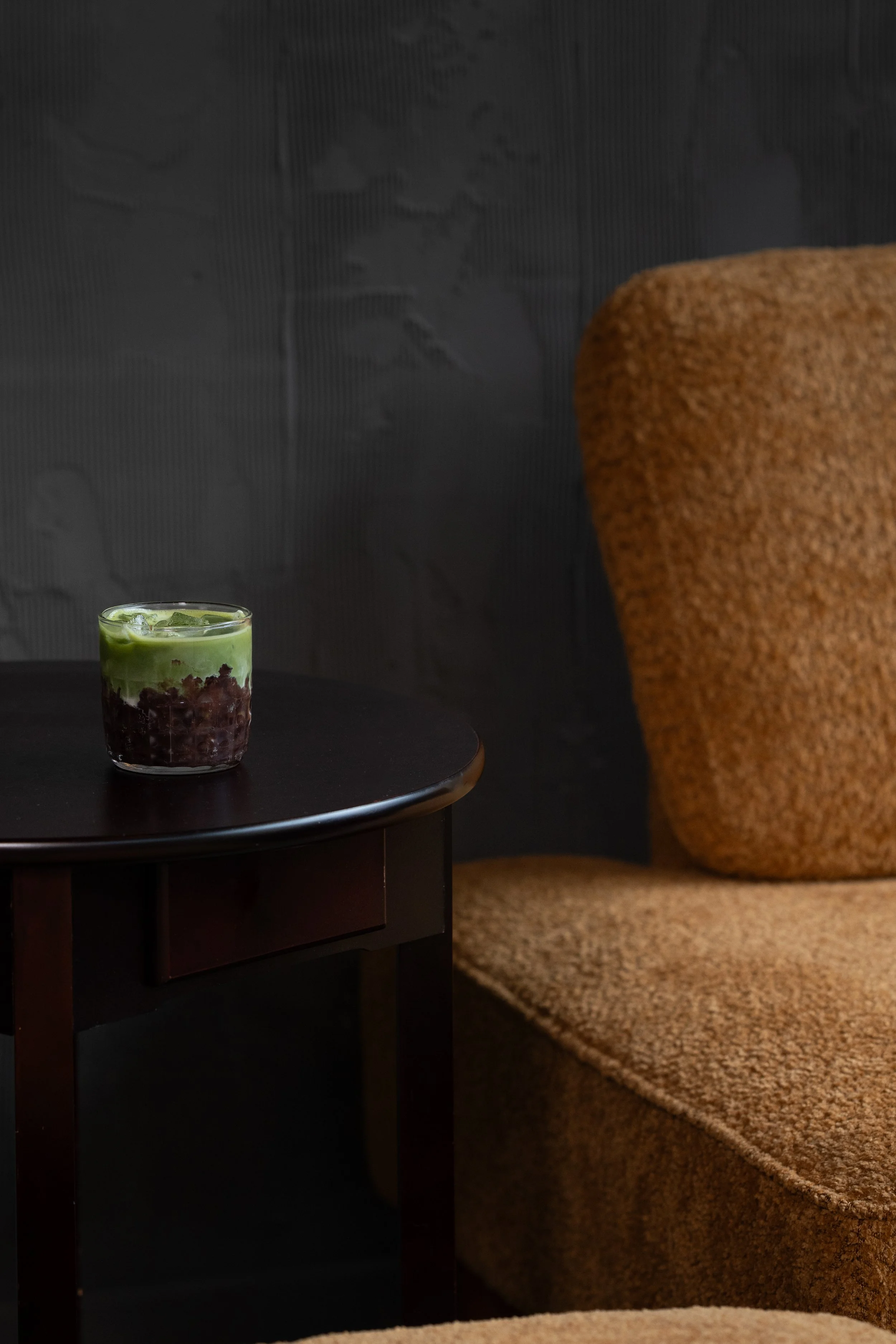 A glass of iced matcha tea on a black side table next to a textured orange-brown armchair in a dark room.