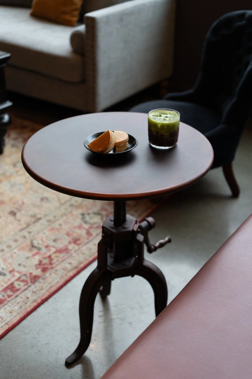 A small round table with a metal base, holding a black plate with a slice of orange and a scoop of ice cream, and a glass of iced green tea, in a cozy indoor setting with a gray sofa and patterned rug.