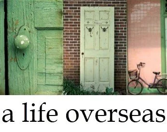 A green wooden door with a vintage green clock on the wall, a white door with black decorative hooks, and a pink bicycle against a pink wall. Text reads 'a life overseas'.