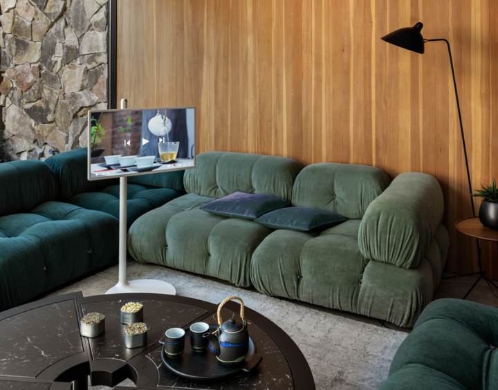 Modern living room with green velvet couches, a wooden paneled wall, a stone accent wall, a black floor lamp, a round black coffee table with candles and a teapot, and a flat-screen TV on a stand displaying a dinner scene.