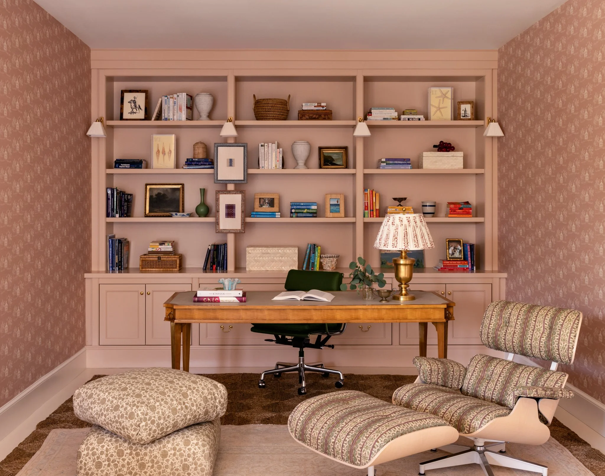 A cozy home office with a built-in pink bookcase filled with books and decorative items. There is a wooden desk with an open notebook, books, a green chair, and a table lamp. The room has pink patterned wallpaper on the walls and a brown carpet, with