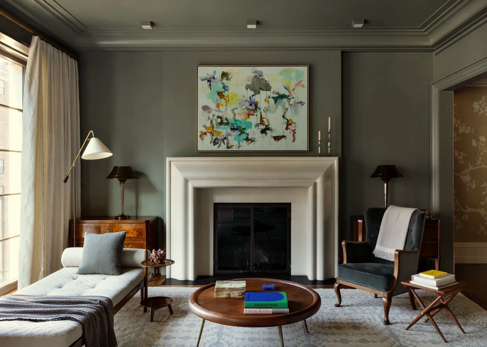 Living room with gray walls, a white fireplace, and an abstract painting above the mantel. There are lamps on each side of the fireplace, a cream-colored chaise lounge, and a dark blue armchair. A round wooden coffee table with books and a decorative