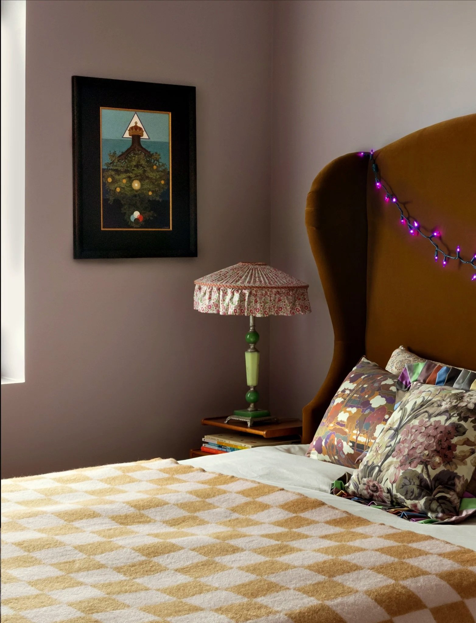 A cozy bedroom corner with a bed, pillows with floral and geometric patterns, a beige and white checkered blanket, a nightstand with a colorful lamp, a framed artwork of a tree and a triangle, and purple string lights on a mustard-colored headboard.