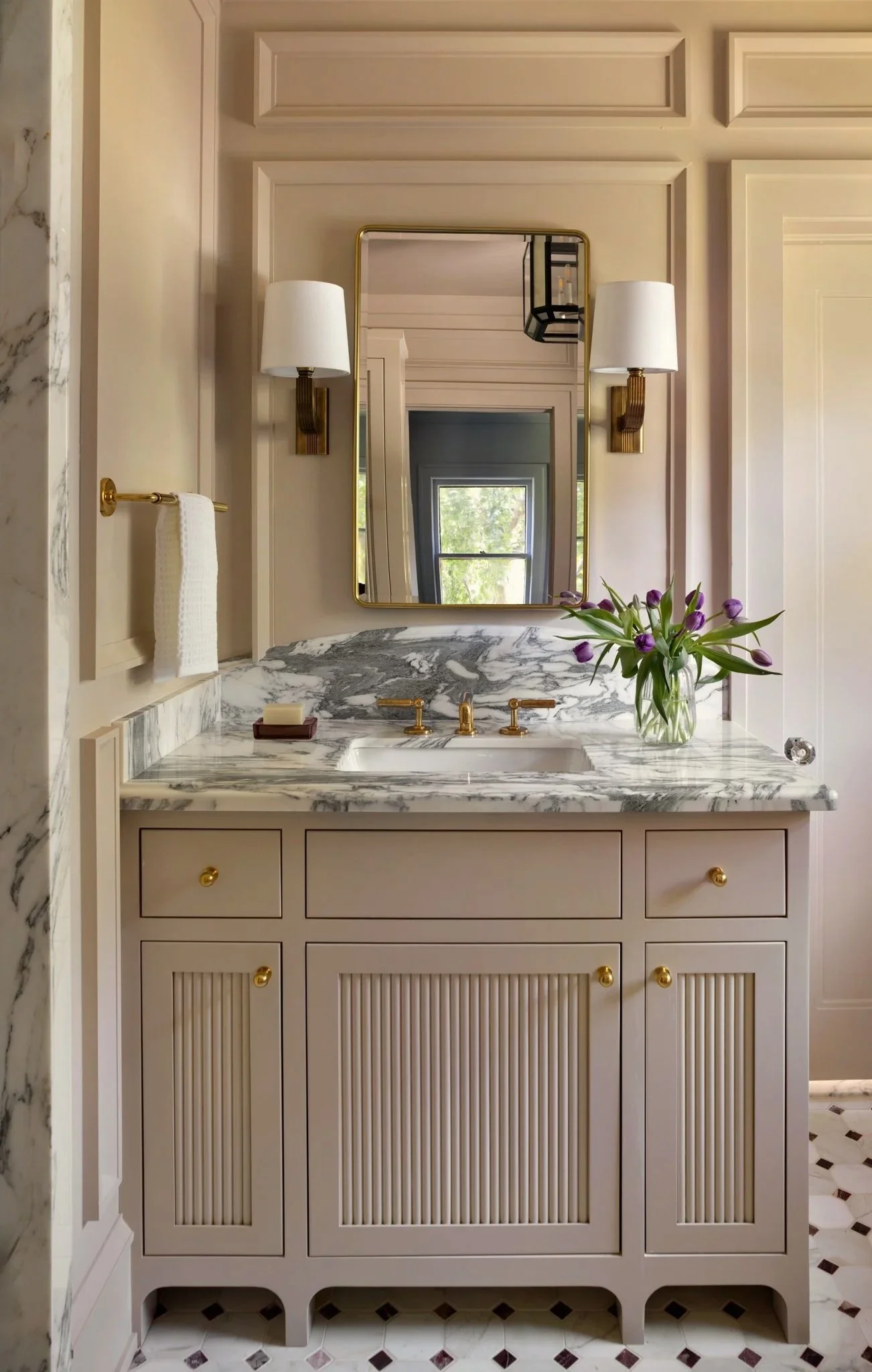 A bathroom vanity with a marble top, gold fixtures, a mirror, and a vase of purple tulips. The cabinet has gold knobs and a ribbed design on the doors, set against a wall with decorative molding and two wall sconces.