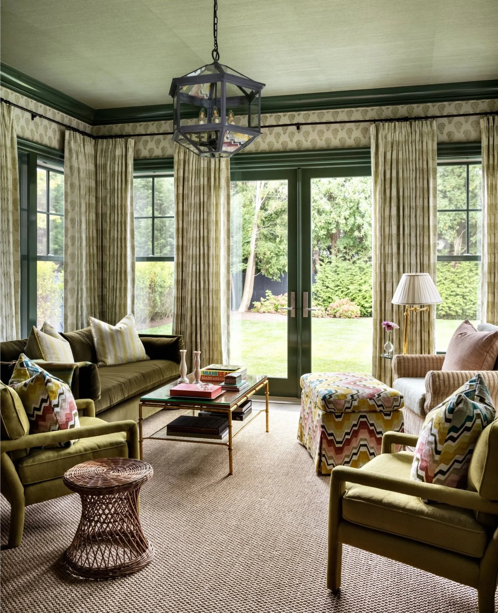 Stylish living room with green sofas, patterned armchairs, wicker stool, glass coffee table, large windows with drapes, and a hanging square light fixture.