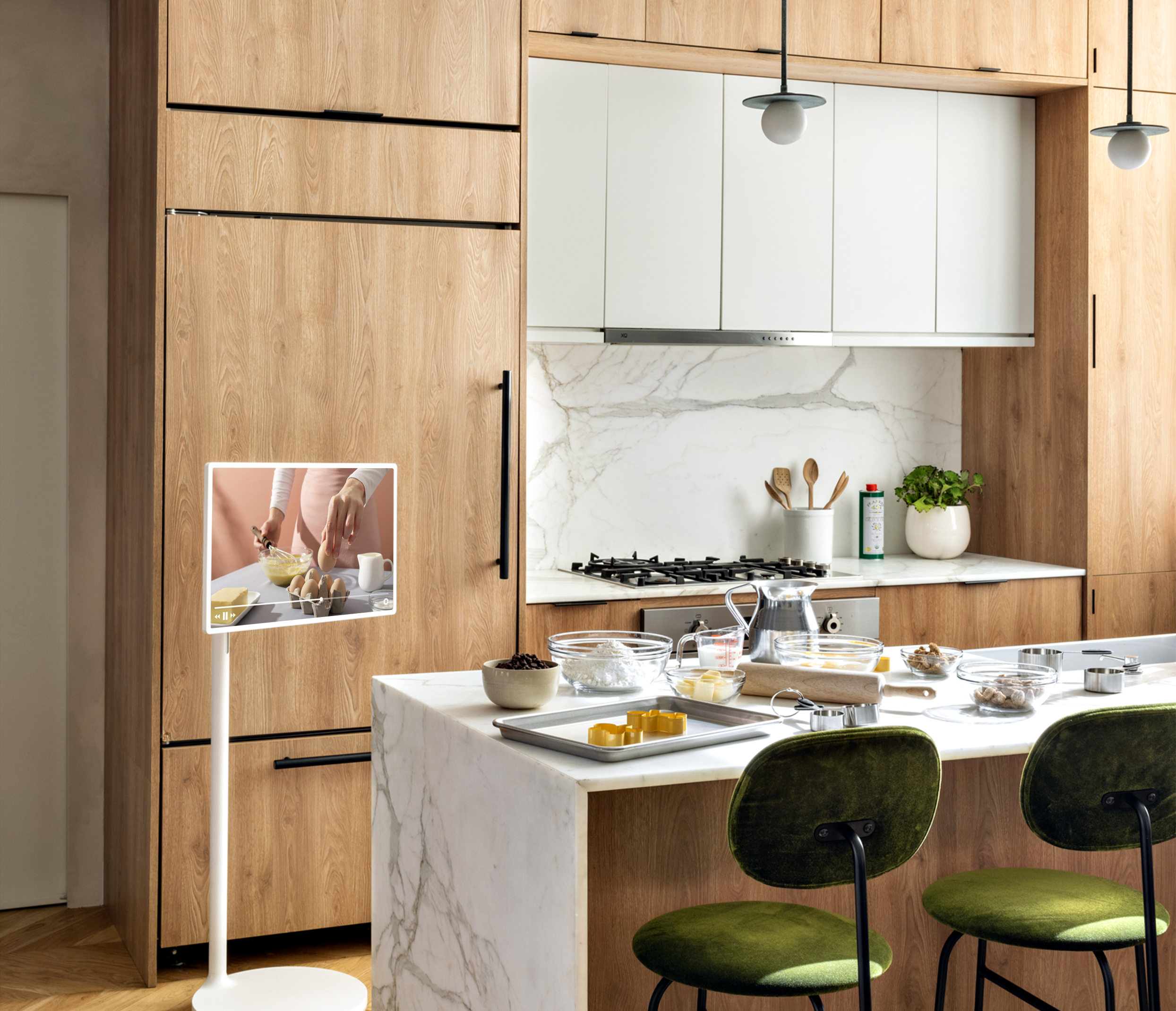 Modern kitchen with wooden cabinets, white marble countertops, and green bar stools. Kitchen island set for cooking with mixing bowls, measuring cups, and ingredients. Wall-mounted screen showing cooking tutorial. Pendant lights hang from the ceiling