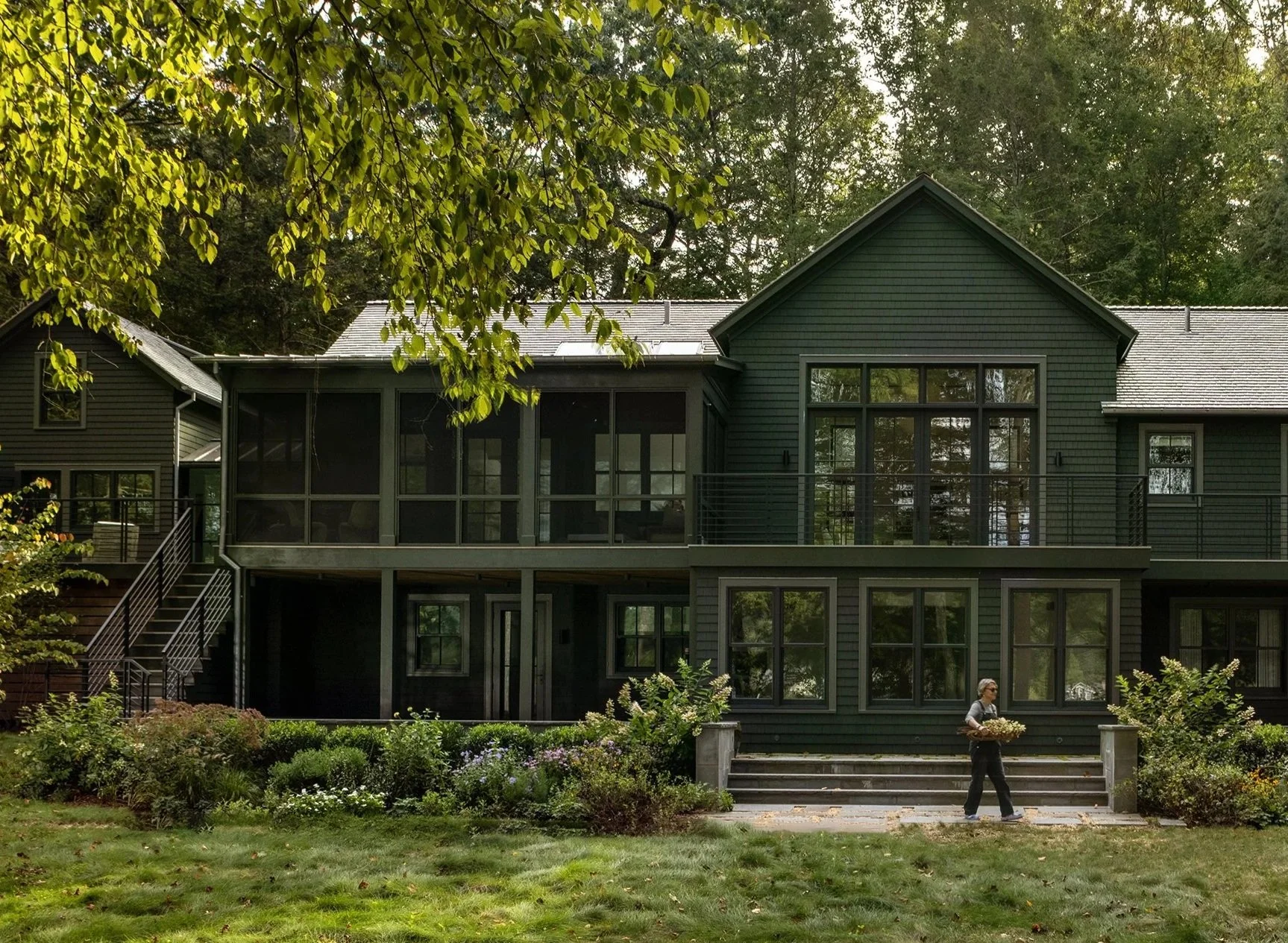A modern dark green house with large windows and a deck is surrounded by lush trees and a well-maintained lawn. A person is walking on the porch carrying a basket of flowers.