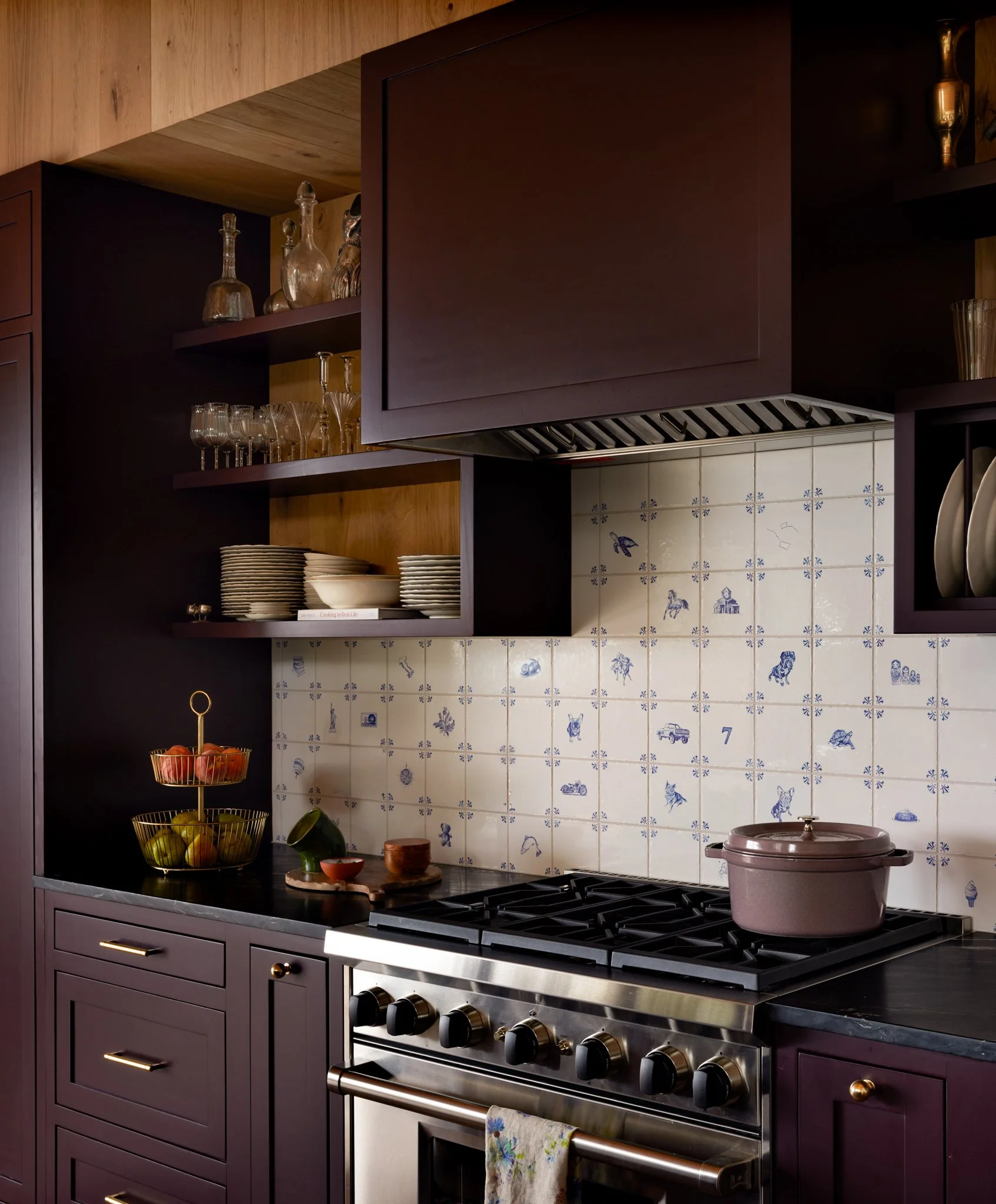 A kitchen with dark purple cabinets, a black countertop, open shelves with glassware and bottles, and a backsplash with blue and white patterned tiles featuring various animals and objects. There is a gold fruit basket with apples and lemons, a pink 