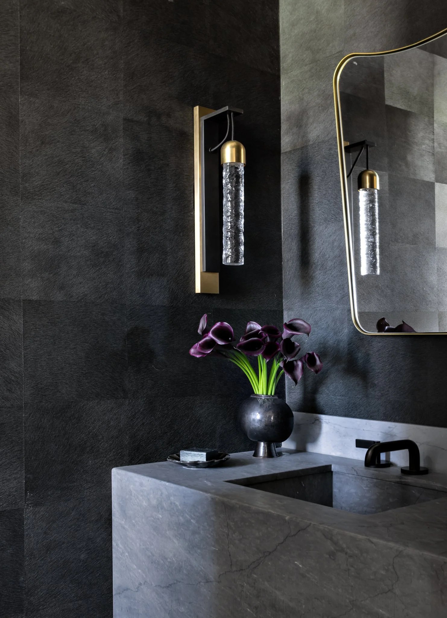 A modern bathroom with dark textured wall tiles, a vanity with a marble countertop, a black vase with purple calla lilies, and a large mirror with a gold frame. Two wall-mounted light fixtures with gold caps and textured glass shades are reflected in