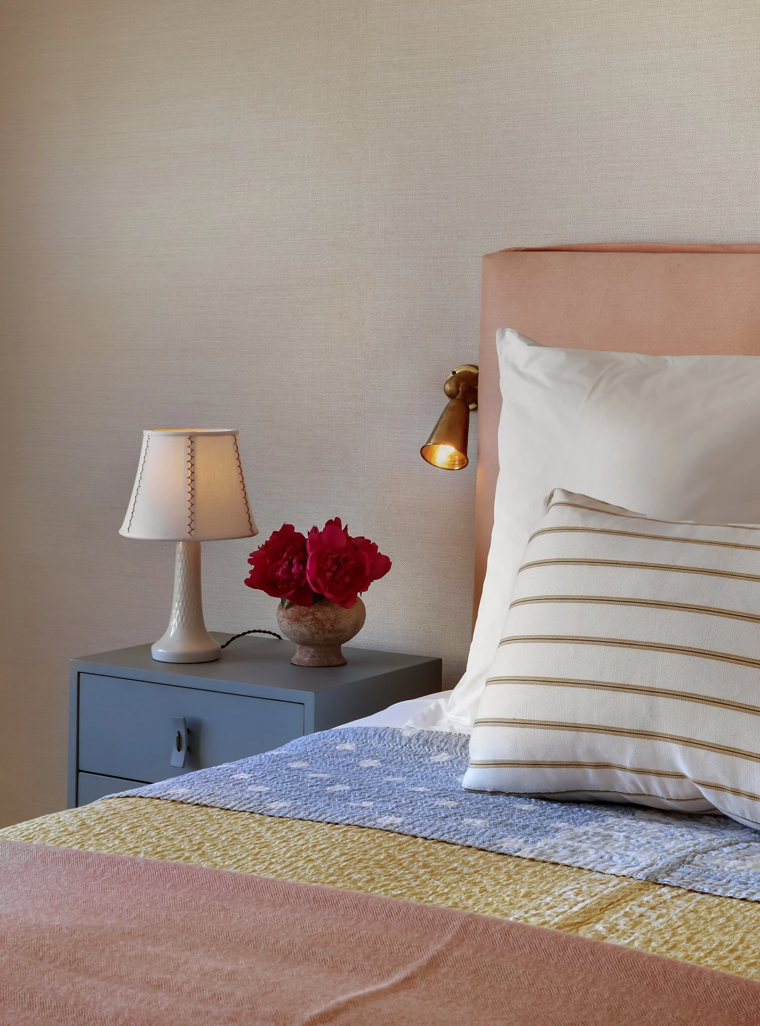 A cozy bedroom with a soft pink headboard, white bed linens with striped pillows, and a matching pink quilt on the bed. There are two bedside lamps, one white with decorative stitching and the other brass, attached to the wall above the nightstand. A