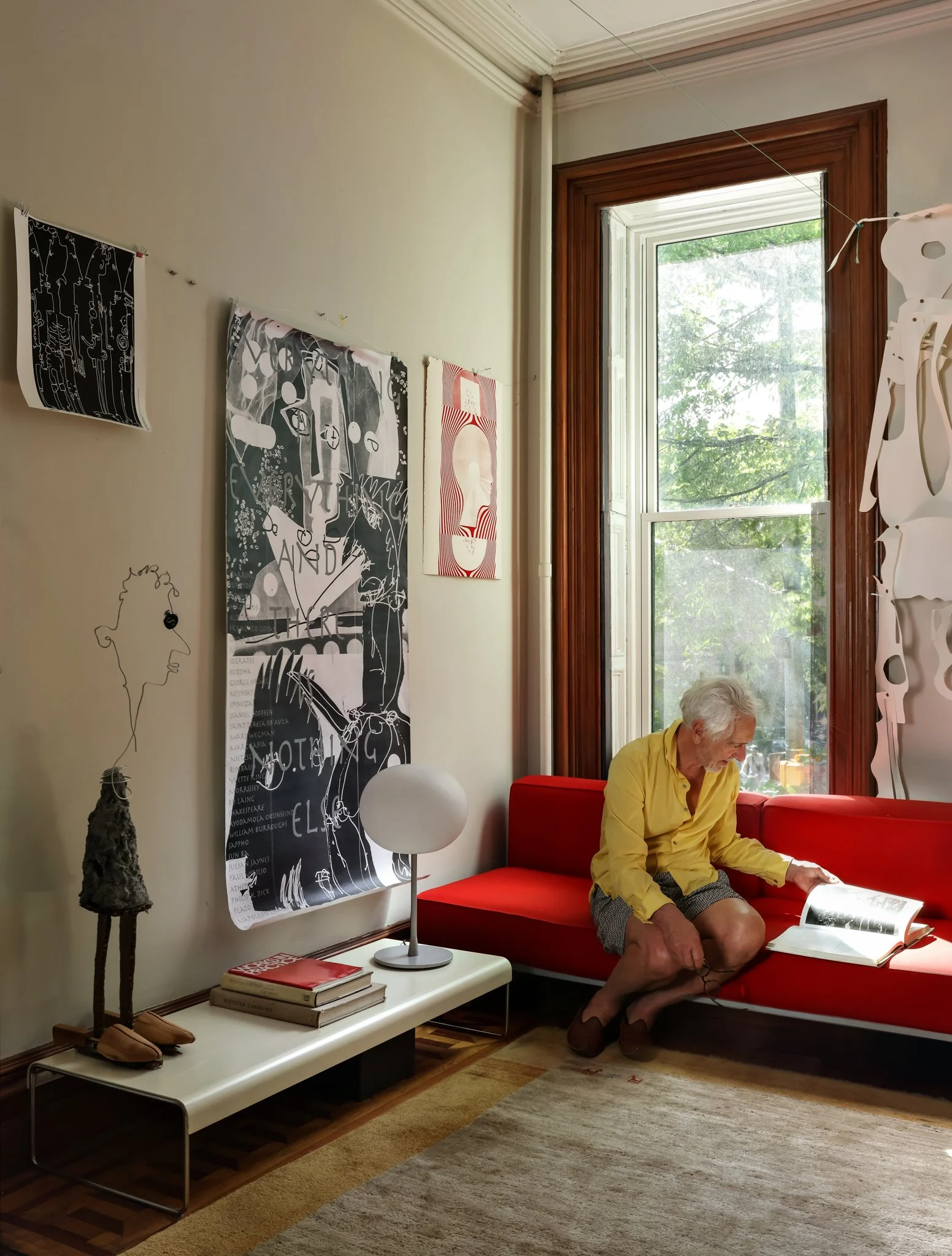 A man with gray hair, wearing a yellow shirt and shorts, sitting on a red sofa, reading a book near a large window with trees outside. The interior has artwork on the wall, a small side table with books and a modern lamp, and a textured area rug.