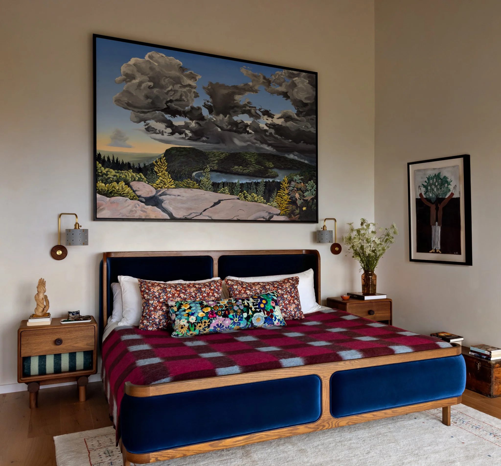 A bedroom with a bed that has a blue upholstered headboard, a red and black checkered blanket, and colorful patterned pillows. There are two wooden nightstands with lamps on either side of the bed. A large landscape painting of clouds over hills hang