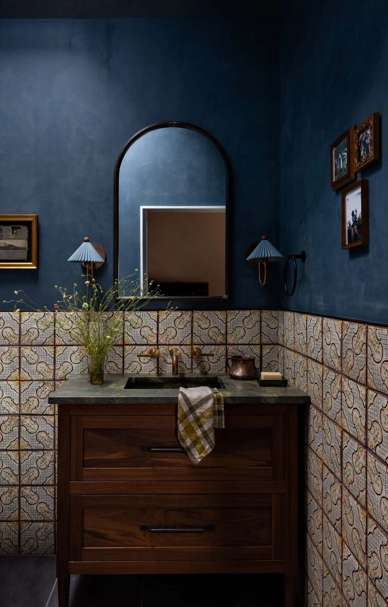 A bathroom interior with a blue painted wall, framed pictures, a mirror, wall-mounted lamps, a sink with a wooden cabinet, a vase with greenery, a bar of soap, and a hand towel.