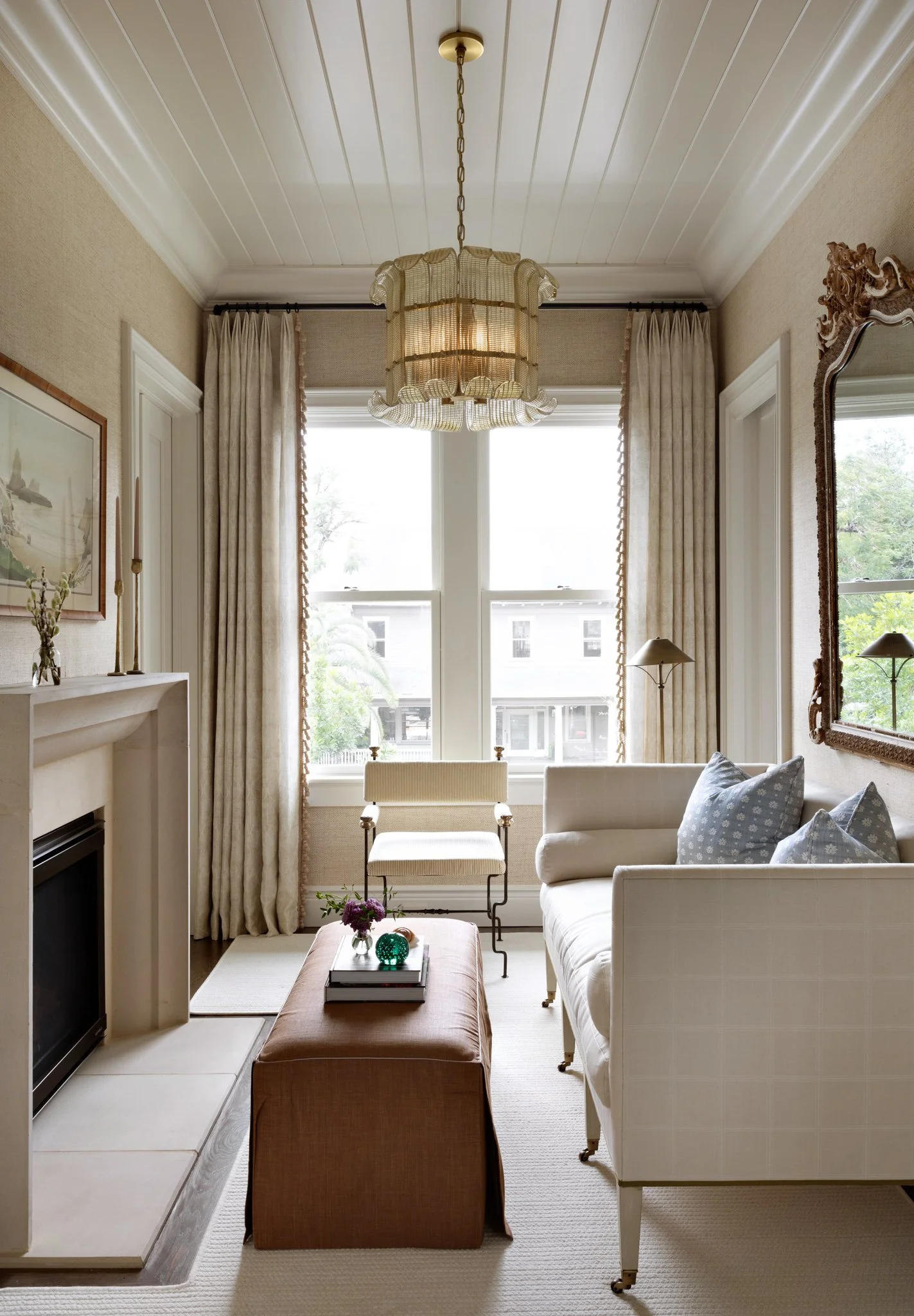 Living room with beige walls and white ceiling, large window with cream-colored curtains, a white sofa with blue pillows, a brown ottoman with decorative objects, a fireplace, a framed picture on the wall, a mirror, and a chandelier hanging from the 