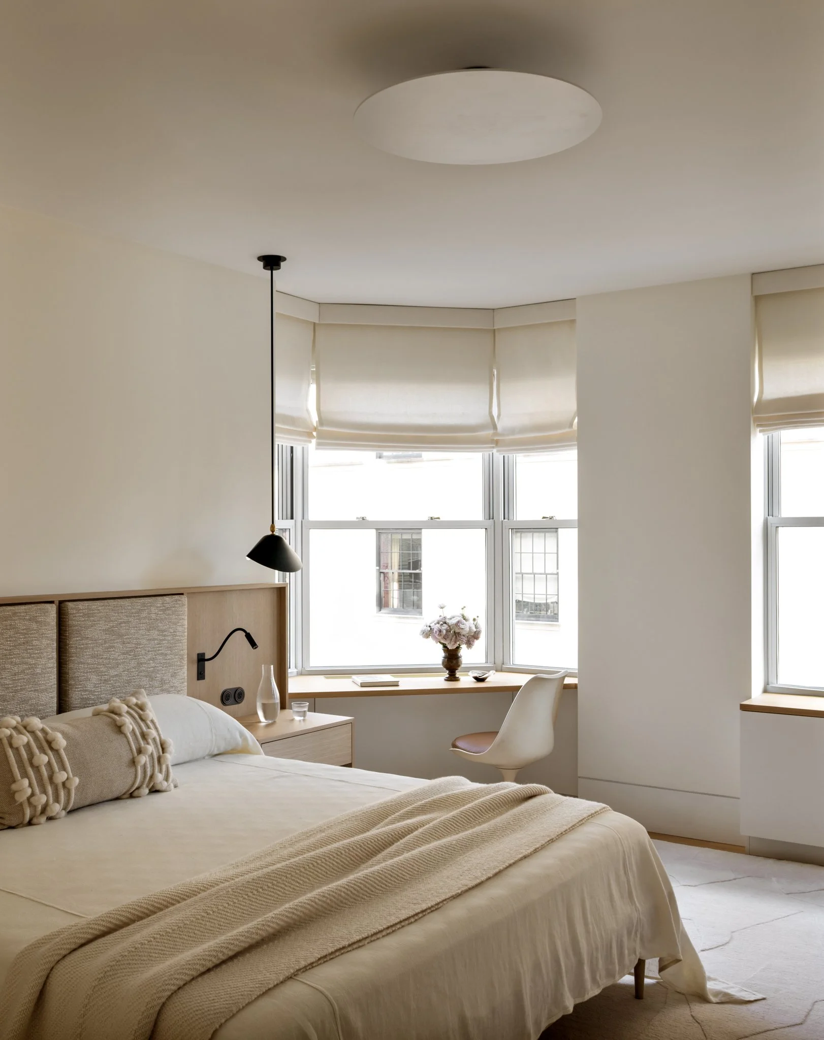 A bedroom with a large bed, beige bedding, decorative pillows, a bedside table with a water bottle, a lamp, and a window with white blinds and a floral arrangement on the windowsill. The room has natural light and neutral-colored walls.