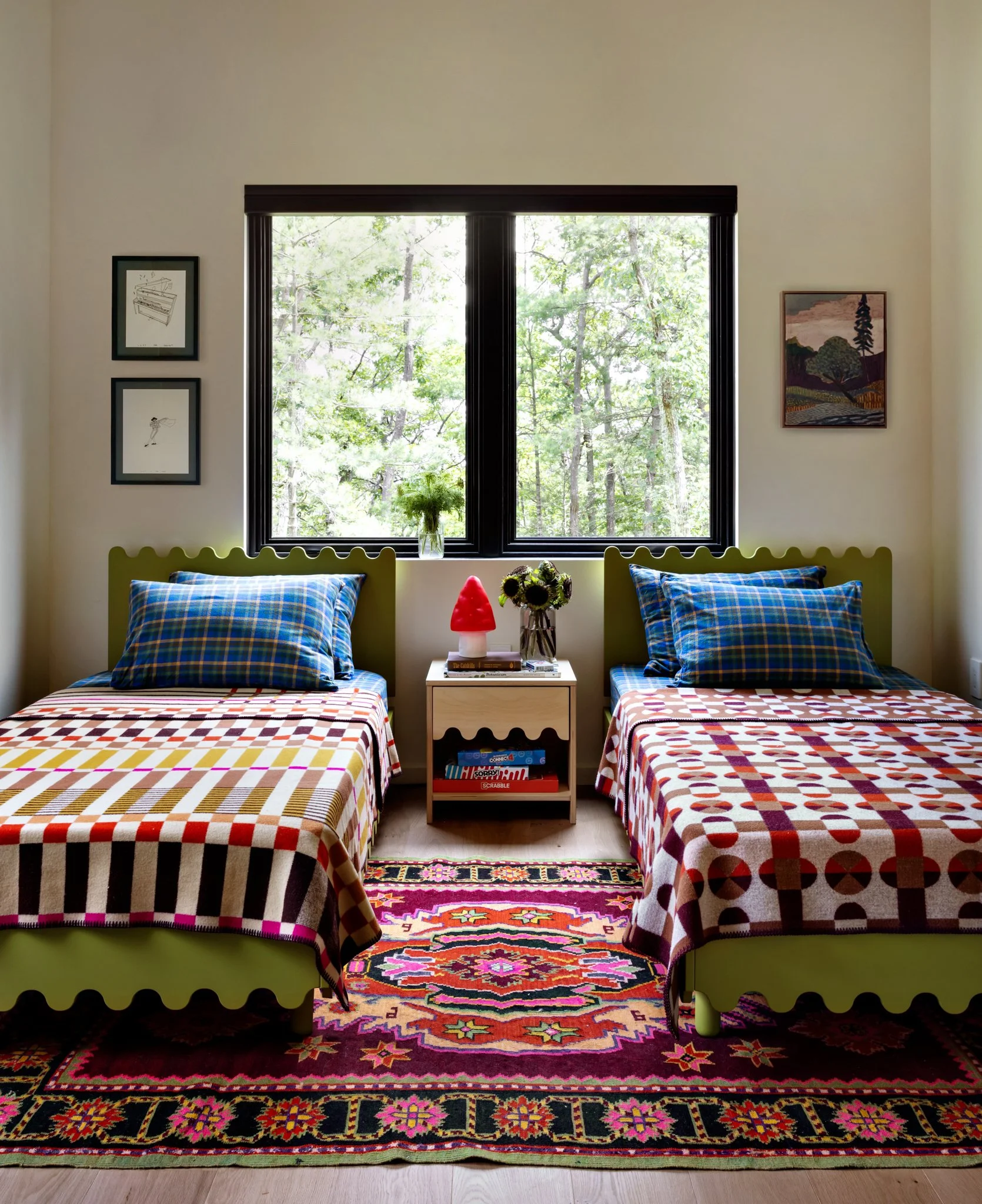 Two twin beds with colorful plaid and geometric patterned bedspreads, positioned side by side in front of a large window overlooking trees. Between the beds is a small nightstand with a red lamp, books, and a vase of flowers. The wall above the beds 
