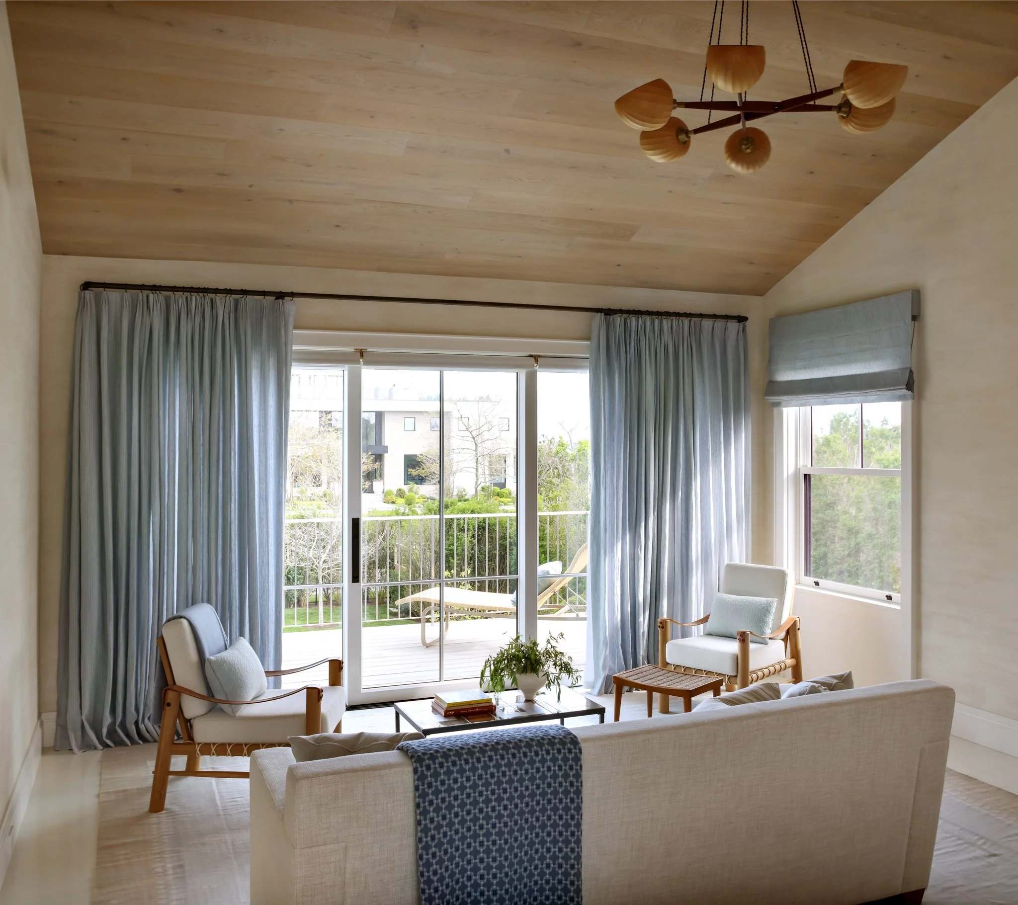 Living room with large windows, white furniture, blue patterned throw, wooden chairs, and a view of the garden outside.