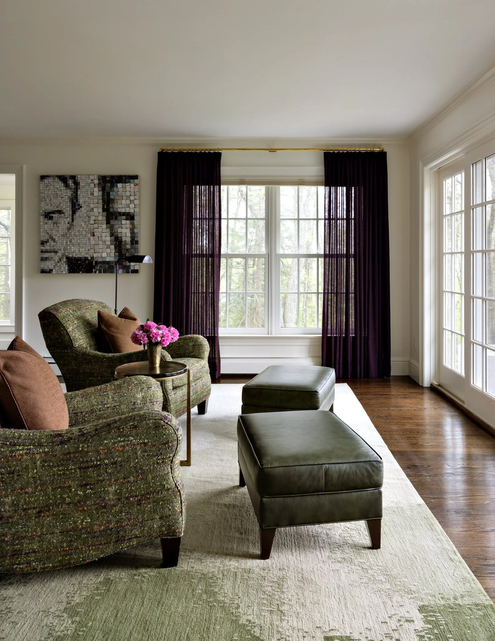 Living room with a large window, dark purple curtains, and a green upholstered armchair with brown pillows, a black ottoman, a small round side table with pink flowers, and a white area rug.