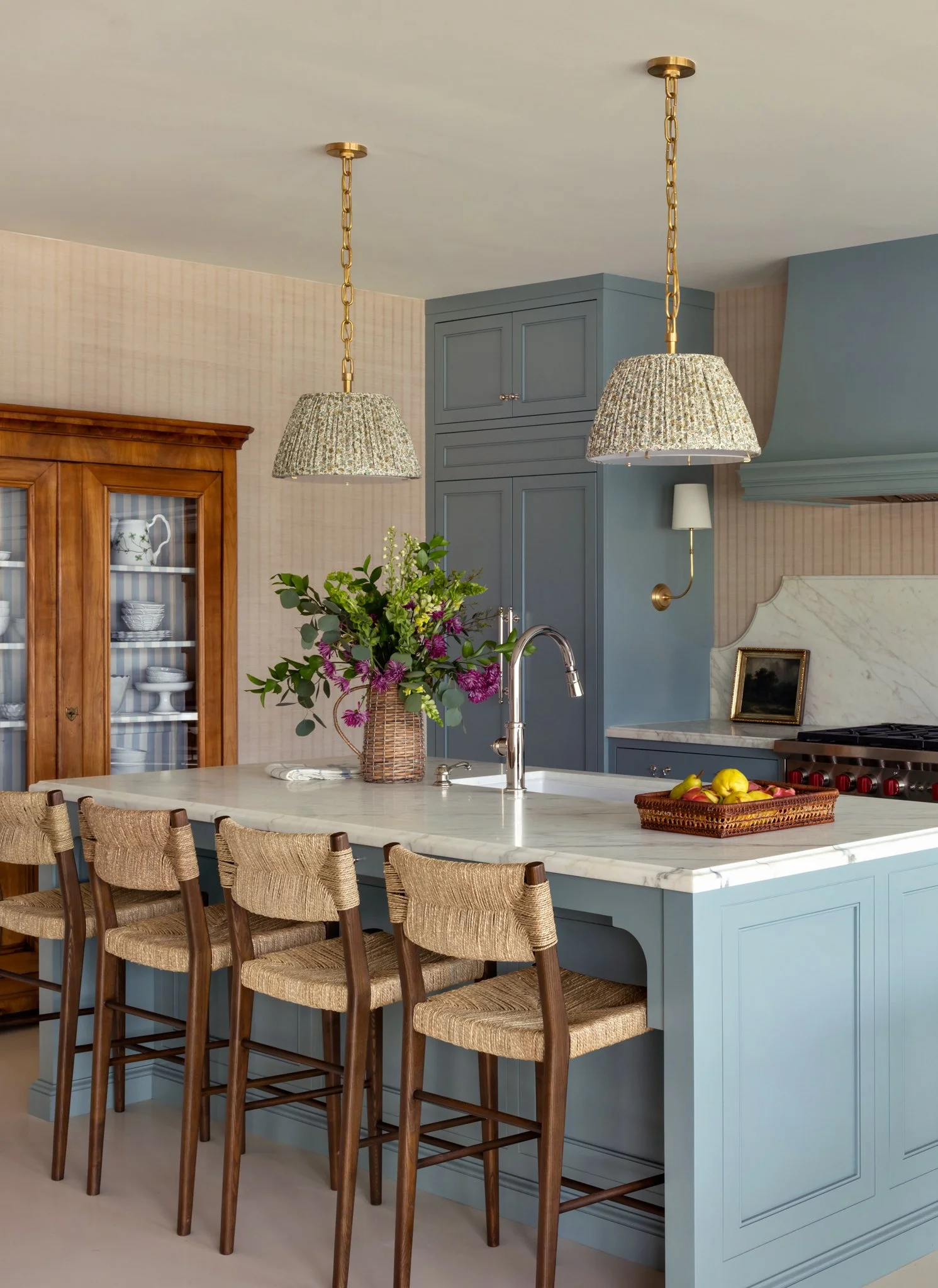 A kitchen with a blue island, wooden bar stools, hanging lamps, a vase of flowers, and a marble countertop.