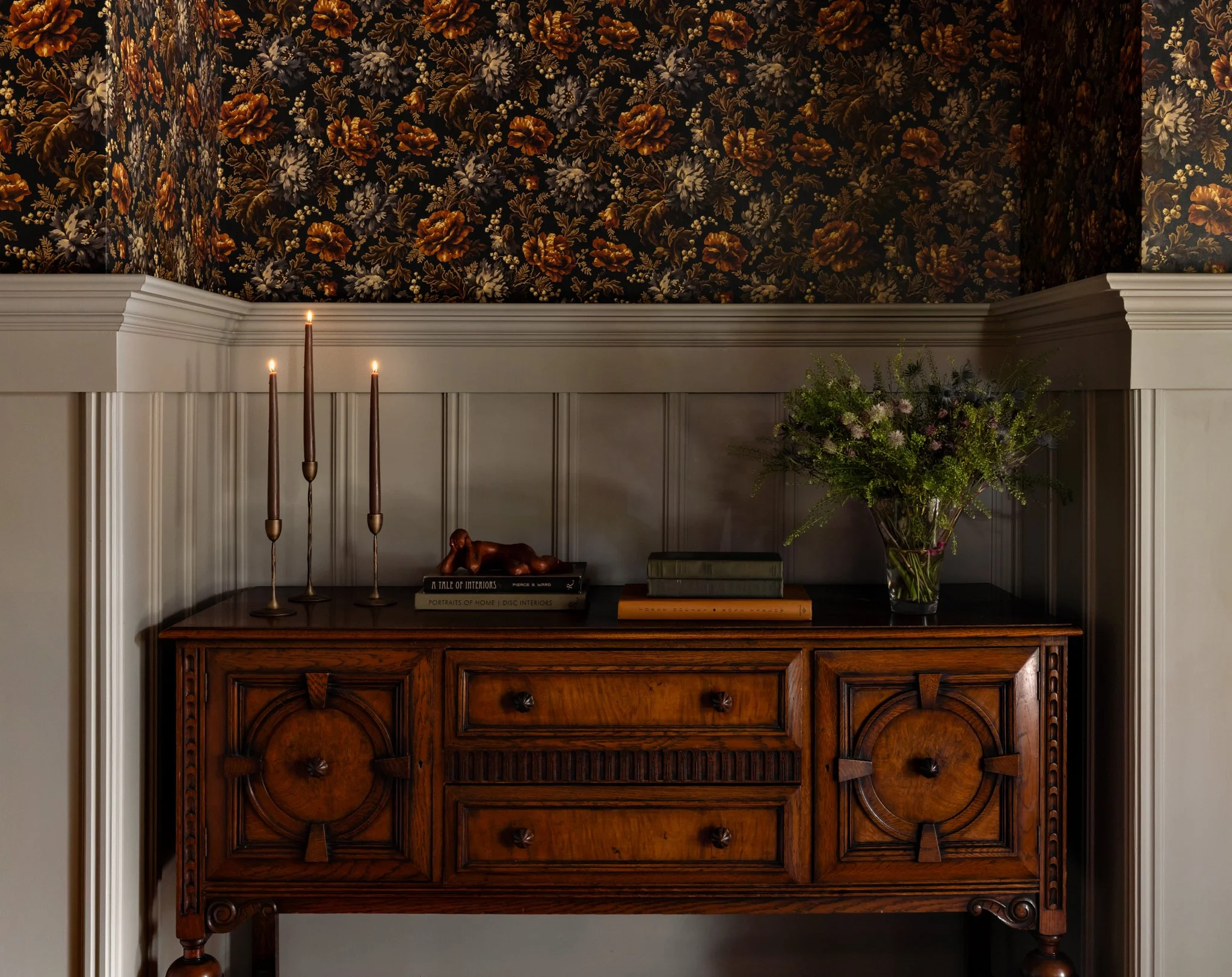 An interior scene featuring a wooden hutch with decorative carvings, three tall candles in holders, a small wooden dog figurine, a stack of books, floral wallpaper and flowers photographed by AD 100 NYC interiors hospitality photographer regan wood