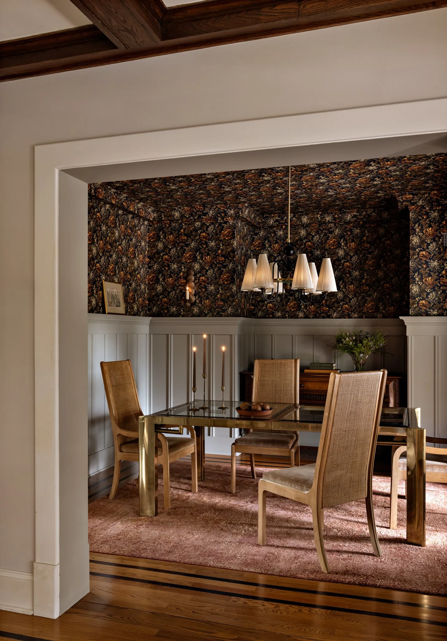 A dining room with a glass-top dining table surrounded by six beige upholstered chairs. There are candles on the table and artwork on the back wall. A six-armed chandelier with shade covers hangs above the table. The room has a pinkish rug, wood floo