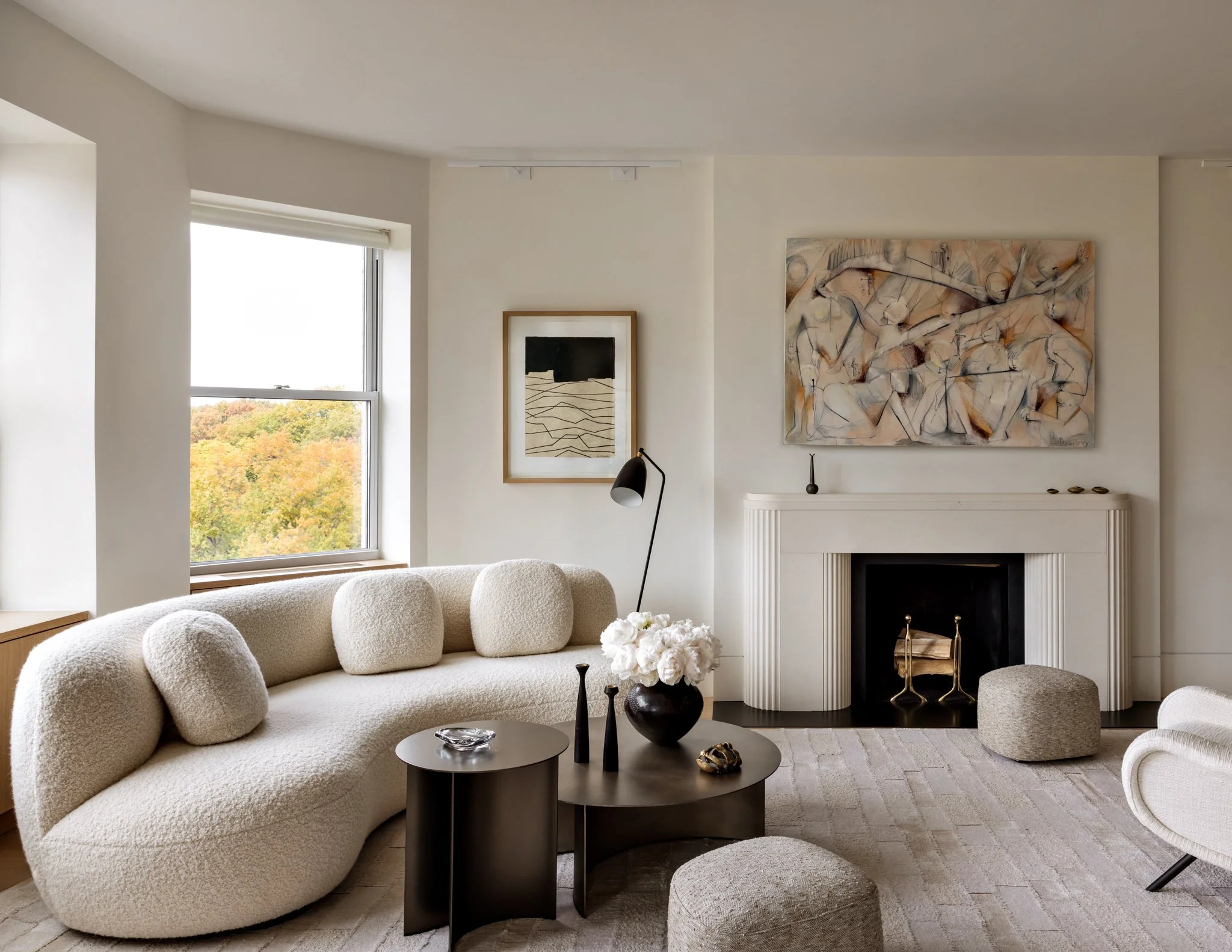 Modern living room with beige textured sofa, round black and beige coffee tables, contemporary art on walls, large window with view of trees, and neutral decor accents.
