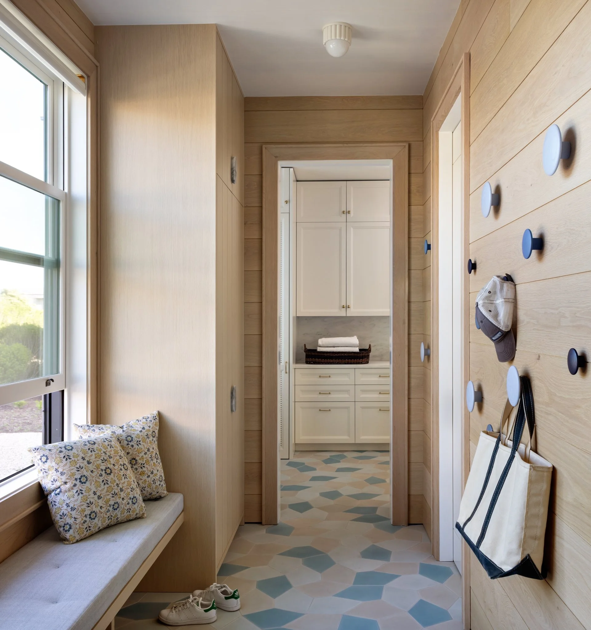 Entryway with wooden walls and floors, a window with a cushioned bench and pillows, white shoes near the bench, and a hallway with white cabinetry and tile flooring. There are hooks with tote bags hanging on the right side.