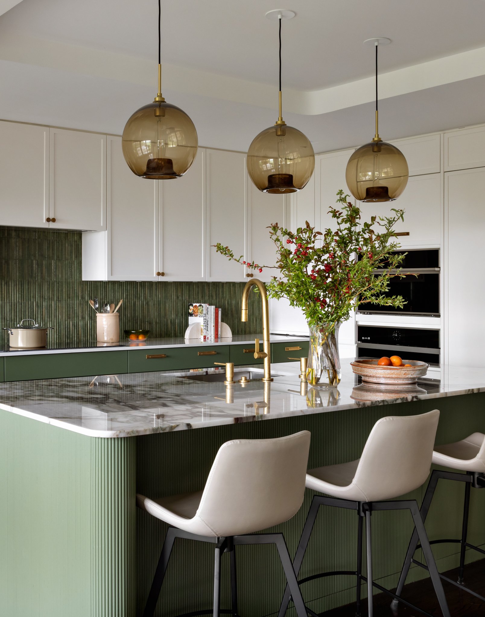 Modern kitchen with white cabinets, green island with ribbed panels, marble countertop, three pendant lights, a vase with greenery, a bowl of oranges, and kitchen appliances.