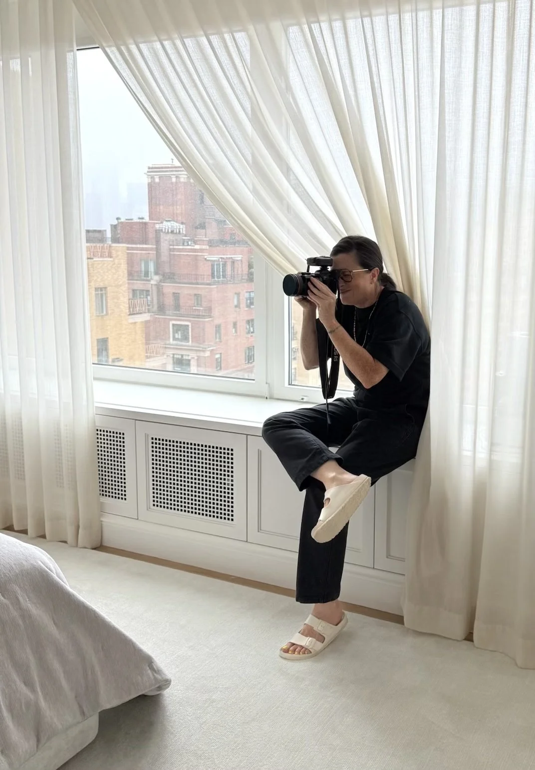 NYC interiors hospitality and lifestyle photographer Regan Wood sitting on a windowsill, taking a photograph with a camera, in a room with white curtains and a cityscape skyline outside.