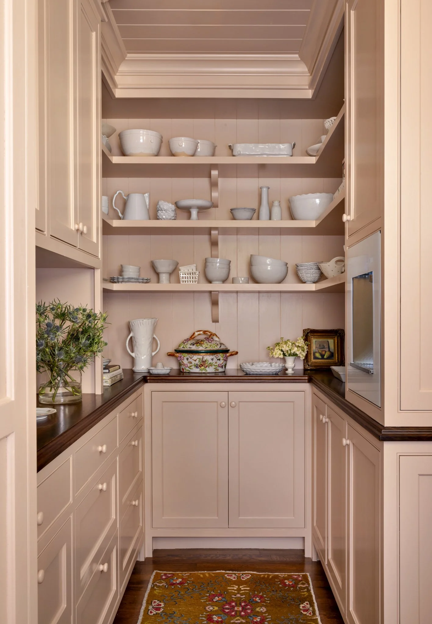 A cozy corner kitchen with beige cabinets, open shelving displaying white ceramic dishes, a dark wooden countertop, decorative flowers, and a colorful rug on the floor.