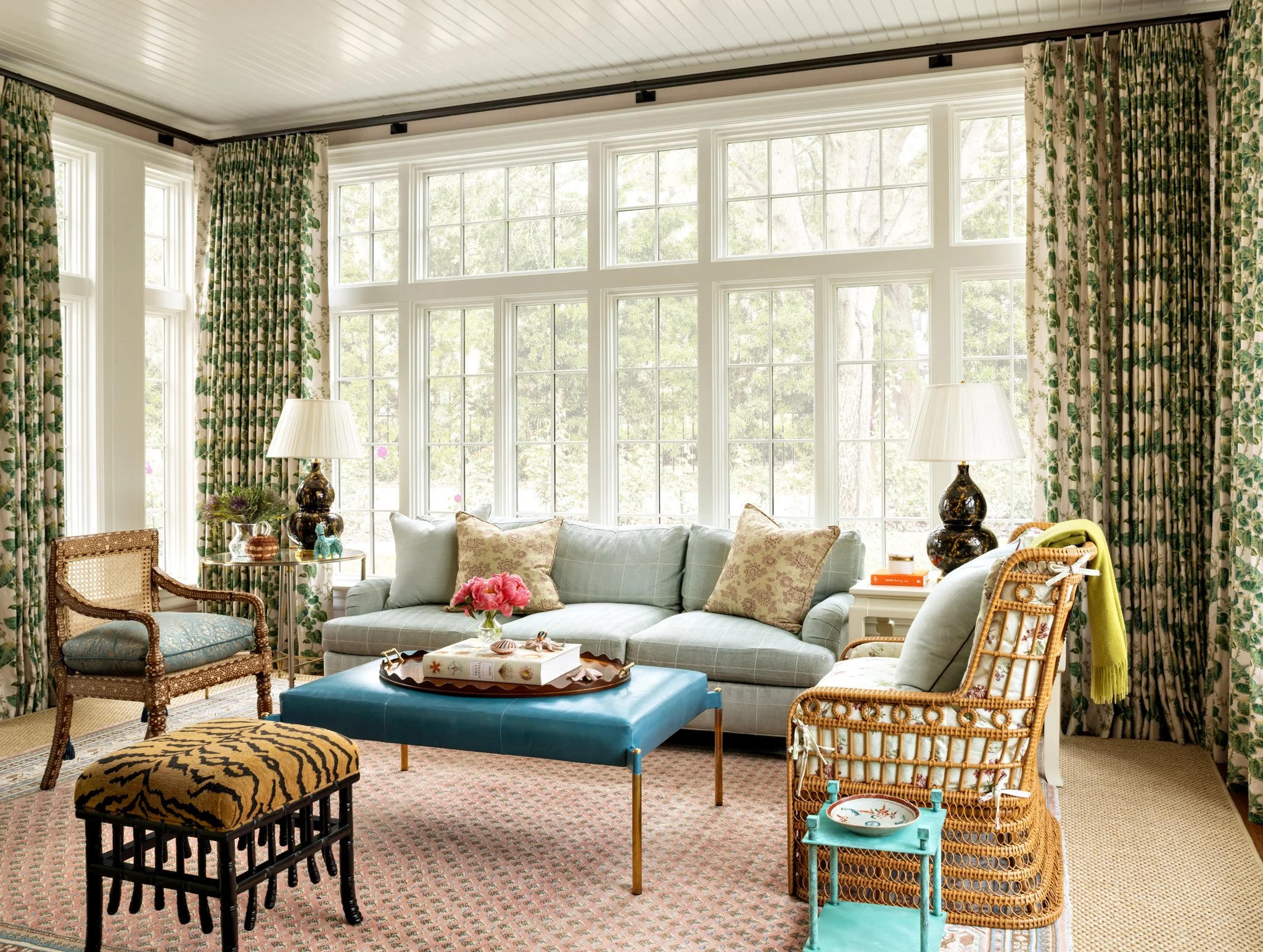 Living room with large windows, patterned curtains, a light-colored sofa with decorative pillows, various chairs, lamps, and a coffee table with a floral arrangement, set on a patterned carpet.