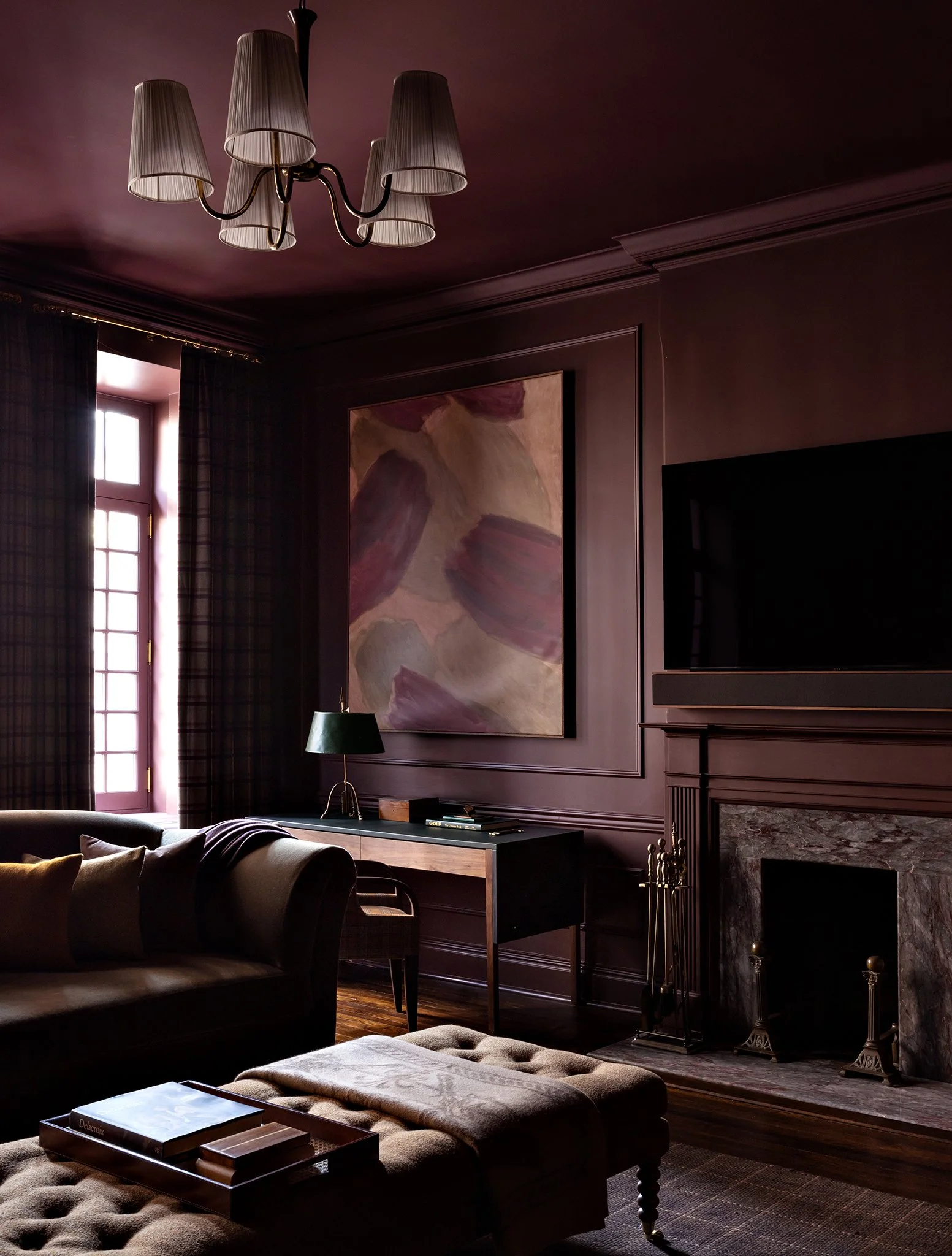 Living room with dark purple walls, a large painting, a fireplace with marble surround, a TV on the wall, a table with a lamp and books, and windows with checkered curtains.